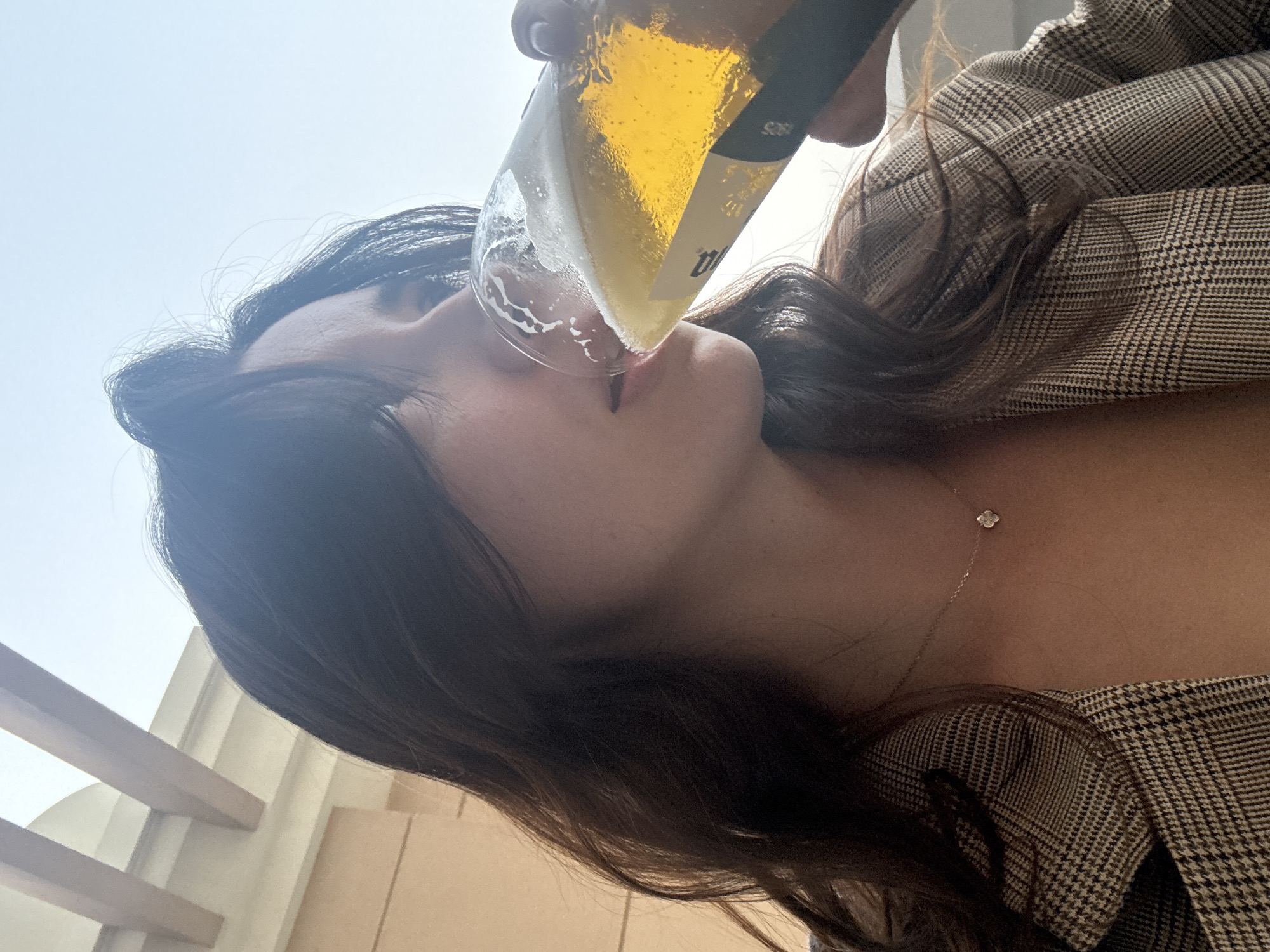 A woman with dark hair is drinking from a bottle, presumably beer. She is wearing a brown and black plaid blazer and a delicate gold necklace with a floral pendant. In the background, there is a bright sky and a partial view of a wooden railing and a light-colored building. It is impossible to definitively confirm the location as Dubai, United Arab Emirates, without additional evidence.