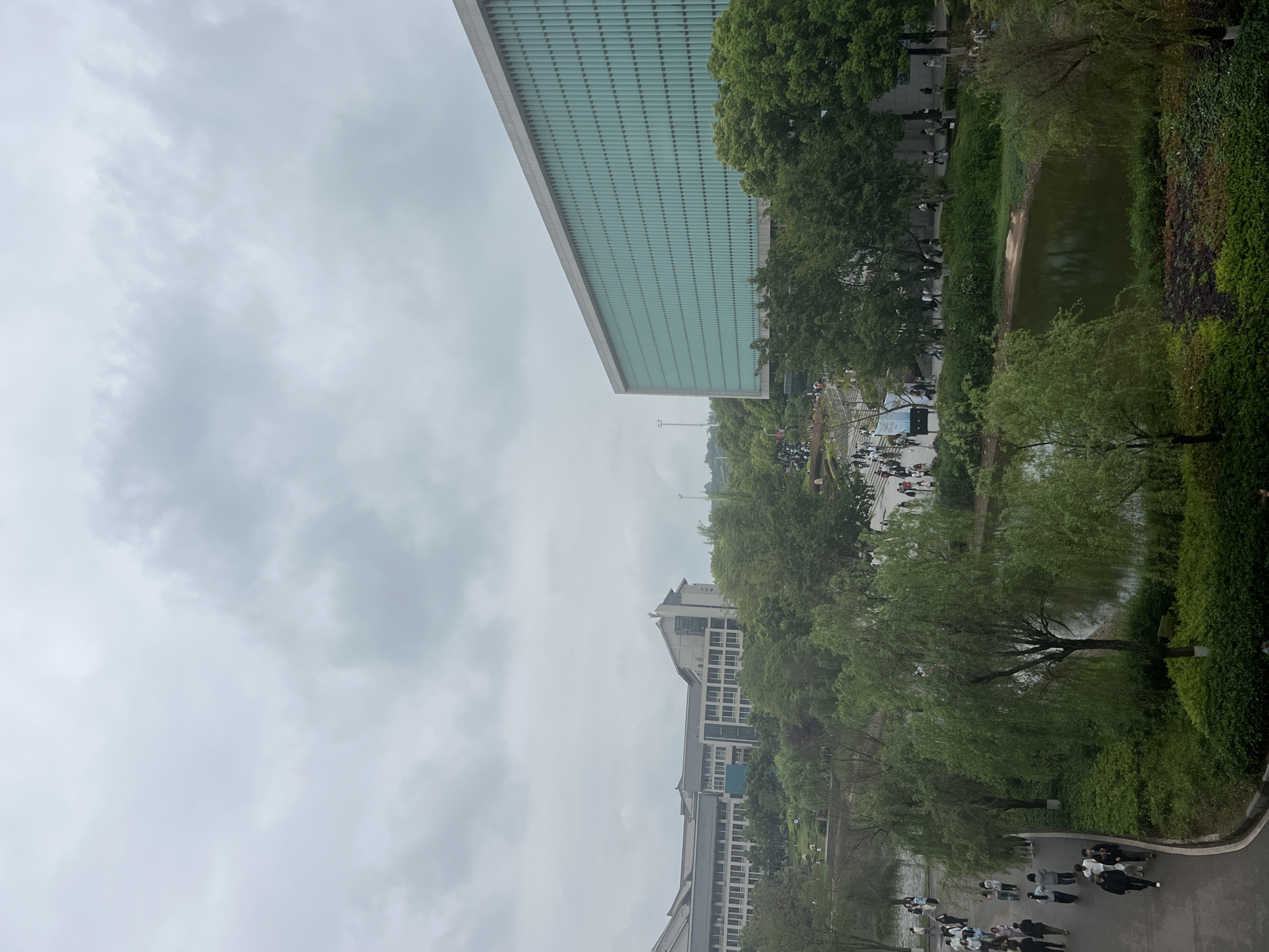 The image shows an overcast day in Jiangjin, China. A large, modern building with a grid-like facade dominates the upper left of the frame. To the right of the building is a narrow body of water, possibly a canal or small lake, with lush green vegetation along its banks. Trees are interspersed throughout the scene, partially obscuring other buildings in the middle distance. A paved pathway runs along the lower portion of the image, where groups of people are walking. In the center of the image, a crowded area suggests a gathering or event. The sky is filled with gray clouds, indicating damp or rainy conditions.