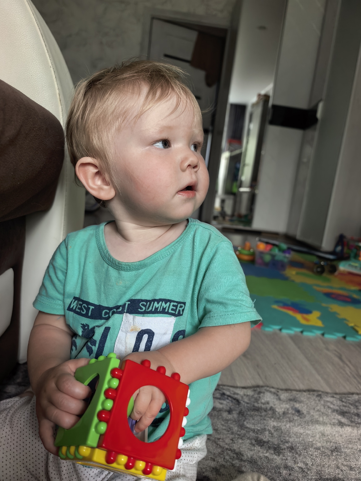 A fair-skinned toddler with blonde hair, wearing a teal t-shirt with the text "WEST COAST SUMMER" printed on it, is sitting on a gray rug and holding a multi-colored toy. The child is looking to the right of the frame, with their mouth slightly open. In the background, a colorful play mat with various shapes and a black toy car can be seen. The scene appears to be an indoor setting, possibly a living room or playroom, with a blurred background that includes furniture and doorways. The image does not provide any information about the city of Sterlitamak, Russia.