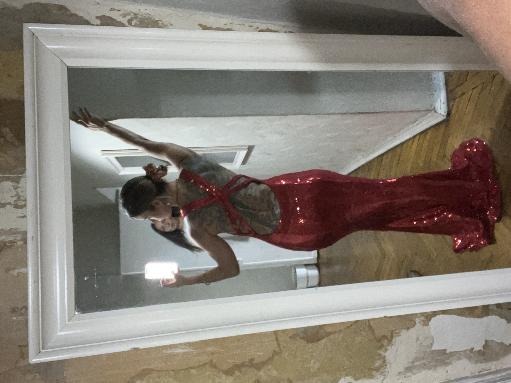 A person is taking a selfie in a full-length mirror. The person is wearing a red sequined dress with a low back and criss-cross straps. They have tattoos on their back and arm, and their hair is pulled up in a bun with a scrunchie. The person is holding a smartphone in their right hand. The mirror is in a room with distressed walls and a wooden floor. The scene appears to be indoors, likely in a home. The lighting suggests it could be daytime, but there is no clear indication of the time of day or weather. The setting looks somewhat unkempt, with peeling paint on the walls. There is no visible text in the image.
