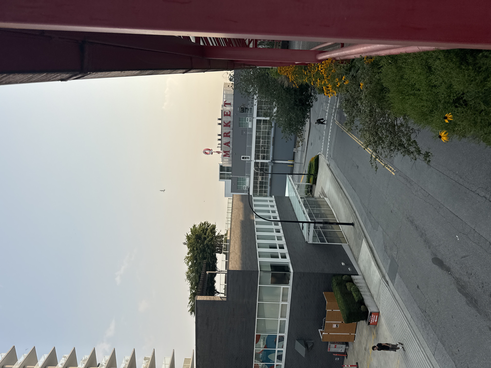In North Vancouver, Canada, a street scene unfolds under a hazy sky. A modern, dark brick building with large windows dominates the left side of the frame, its rooftop featuring trees and a pergola. Across the street, a building with a prominent "MARKET" sign is visible. A person walks on a sidewalk near the brick building, while another person is seen further down the street near a crosswalk. Yellow flowers bloom alongside a red structure that partially frames the right side of the image. The street appears wet in places, indicating recent rain or cleaning.