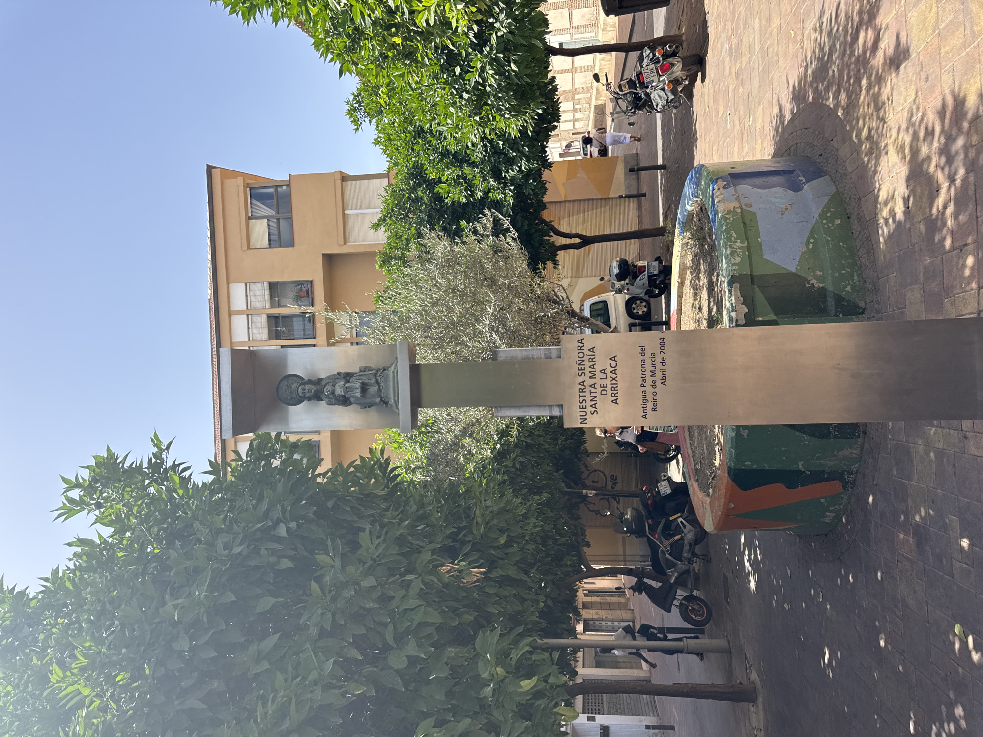 The image shows an outdoor scene in what appears to be a town square in Murcia, Spain. In the foreground, there is a decorative, cylindrical planter with a colorful, abstract design. A metal plaque is mounted on a tall, slender post in front of the planter. The plaque has text written on it, which reads: "NUESTRA SEÑORA SANTA MARIA DE LA ARRIXACA" and below that, "Antigua Patrona del Reino de Murcia, Abril de 2004". This indicates the plaque is a dedication to a patron saint of Murcia.
Behind the planter and plaque, there are several trees, including an olive tree and leafy green trees. A building with an orange facade and balconies is visible in the background. There are also several motorcycles parked on the brick-paved ground. Two people are visible: one man walking away from the camera in the left part of the frame, and a woman in the right part of the frame walking away from the camera. The sky is clear and blue. The overall impression is a sunny day in a public space within a Spanish city.