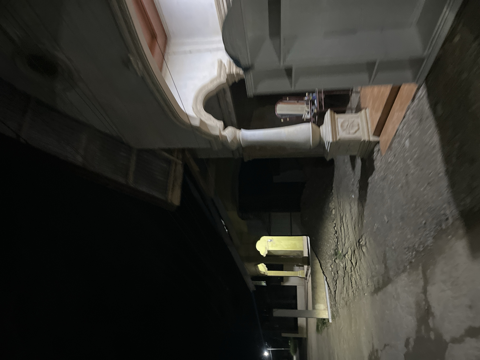 This is an exterior, eye-level, low-angle, nighttime shot of a dimly lit, weathered stone path with some cracks and debris. To the left of the path is a dark, shadowy area, possibly a courtyard or an overhang. In the background, there's a building with arched openings and decorative elements. One of the arches reveals a brightly lit interior, appearing to be a room with yellow walls. The architecture suggests it could be an older, possibly religious or historical building, given the ornamentation on the pillars and the arched construction. The lighting is artificial and uneven, creating strong shadows and highlighting certain architectural features while leaving others in darkness. The overall mood is mysterious and somewhat eerie due to the darkness and the stark contrast between the lit and unlit areas. There is no visible text in the image. The scene appears to be outdoors at night in Menanggini, Indonesia.