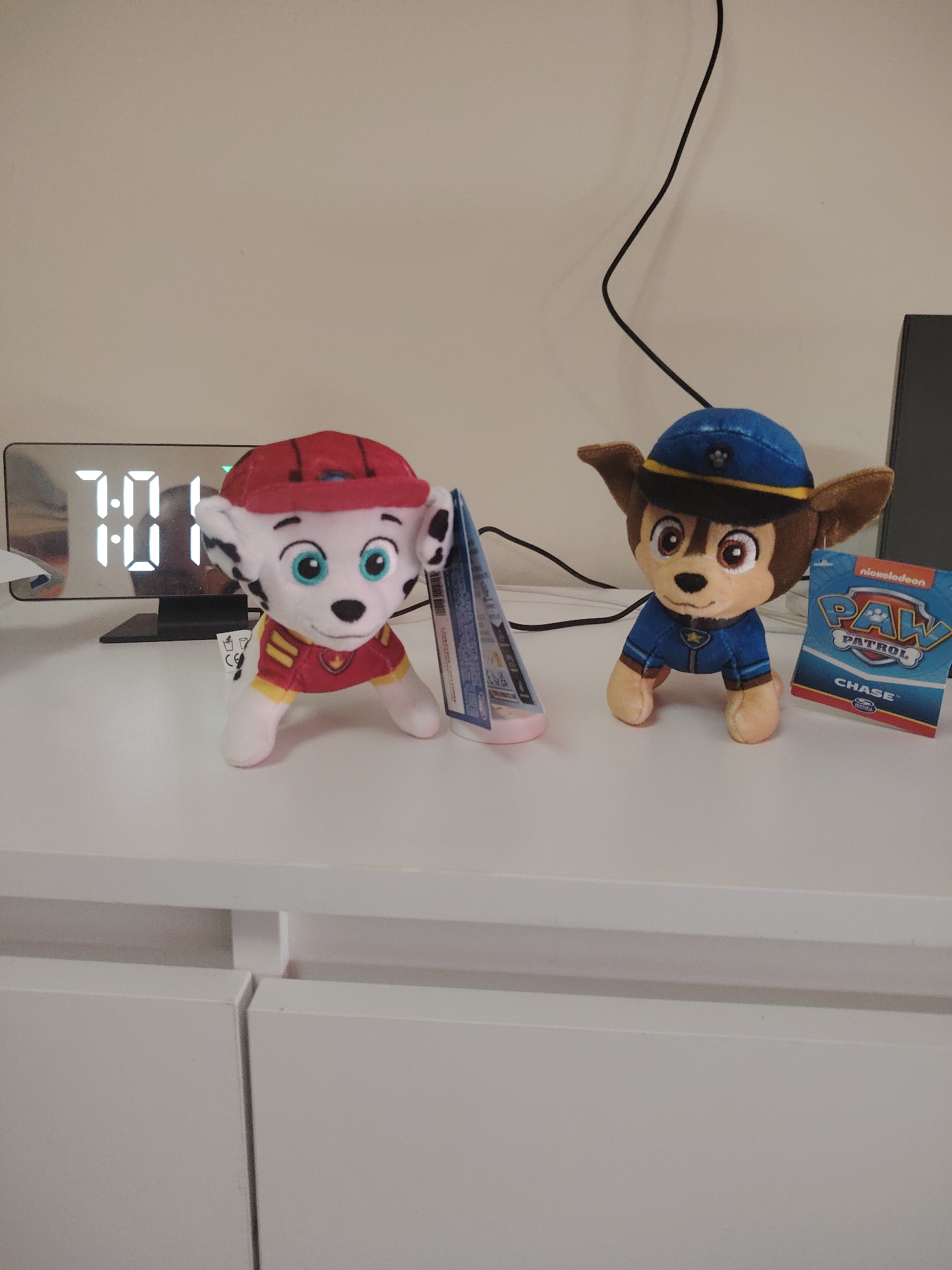The image captures an indoor scene in Huntingdonshire, United Kingdom, featuring two Paw Patrol plush toys displayed on a white surface, likely a desk or shelf. To the left is Marshall, a white plush dog with black spots, wearing a red and yellow firefighter uniform and a red cap. A small white tag with a "CE" logo is visible near his leg. Beside him is a reflective black digital clock displaying "7:01".
To the right, standing beside Marshall, is Chase, a brown plush dog dressed in a blue police uniform and a matching blue police cap. A hang tag is attached to Chase, clearly showing the text "Nickelodeon", "PAW PATROL", "CHASE", and "SPIN MASTER". Between the two plush toys, there's a slender, triangular object with a pink base and partially visible text, which is unreadable. A black cable runs from behind the clock upwards and then curves down behind Chase.
The background consists of a plain light-colored wall. Below the white surface are white cabinets or drawers, indicating a modern, minimalist interior. The lighting suggests artificial light, consistent with an indoor setting, and the digital clock indicates an early morning or evening time. No activity is taking place; the scene is a static display of collectible items.