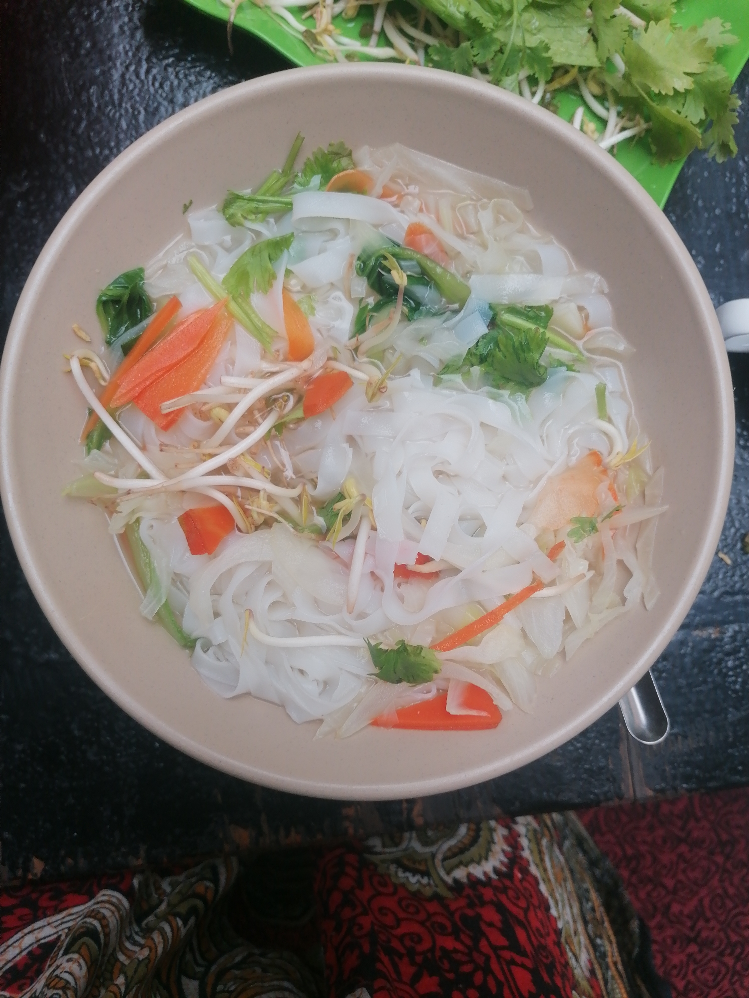 A high-angle shot depicts a bowl of Vietnamese rice noodle soup (possibly Pho) in Bai Chay Ward, Vietnam. The beige bowl is filled with flat rice noodles, thinly sliced carrots, bean sprouts, cilantro, and possibly spinach or another leafy green vegetable, all in a clear broth. A plate with more bean sprouts and cilantro sits in the upper right corner of the image. The bowl rests on a dark, possibly textured surface, and a portion of a patterned fabric, likely clothing, is visible in the lower part of the frame. A spoon is also partially visible beside the bowl.