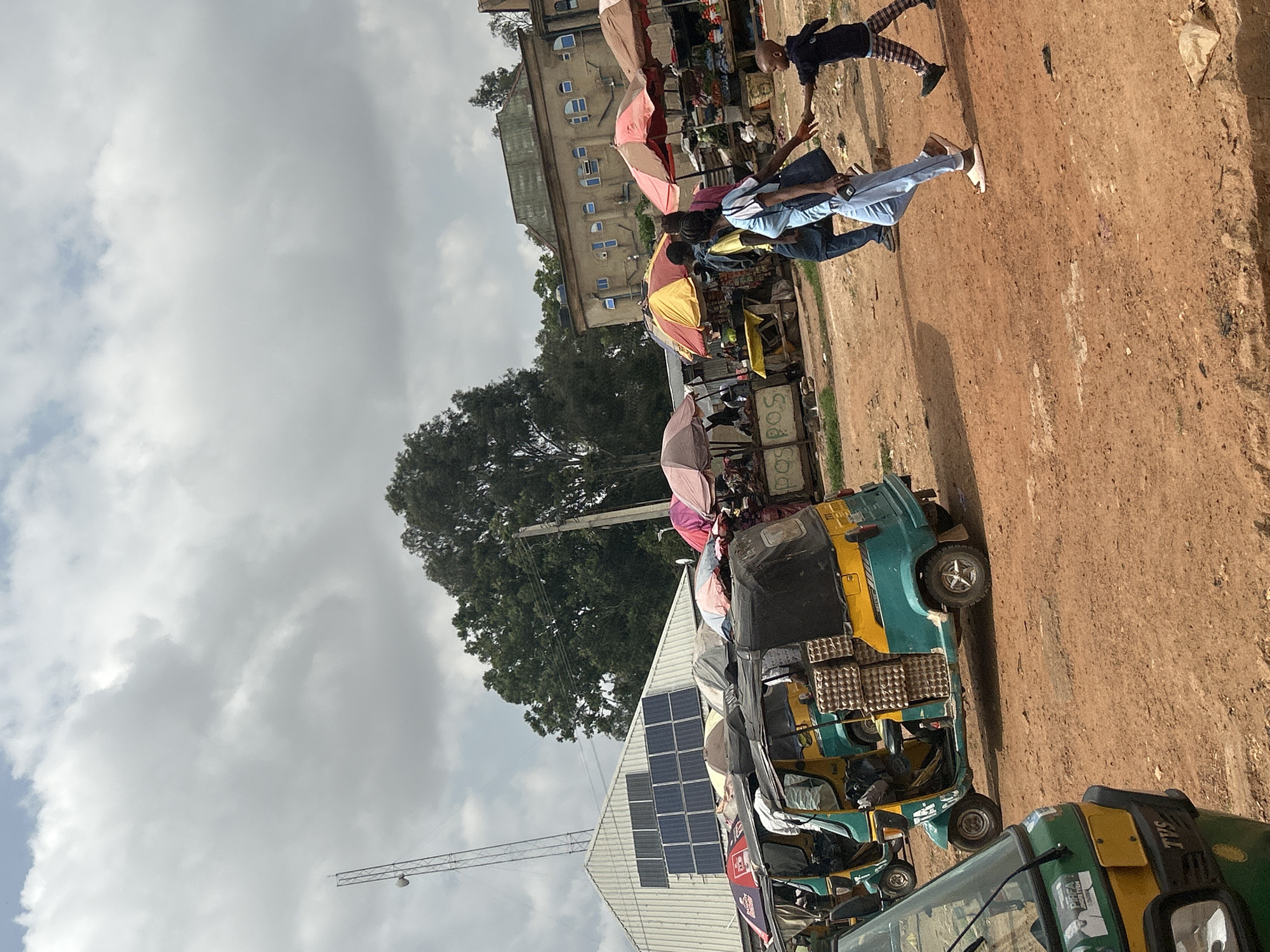 The image shows a street scene in Bar Jirgi Summa, Nigeria. There are several auto rickshaws (Tuk-tuks) parked on the dirt road. Some of the rickshaws have egg cartons stacked on top. Along the road, there are vendors with umbrellas shading their stalls. People are walking and interacting near the stalls. In the background, there is a building with solar panels on its roof. Above the building and rickshaws, there is a large tree. The sky is cloudy. A metal tower is visible.
