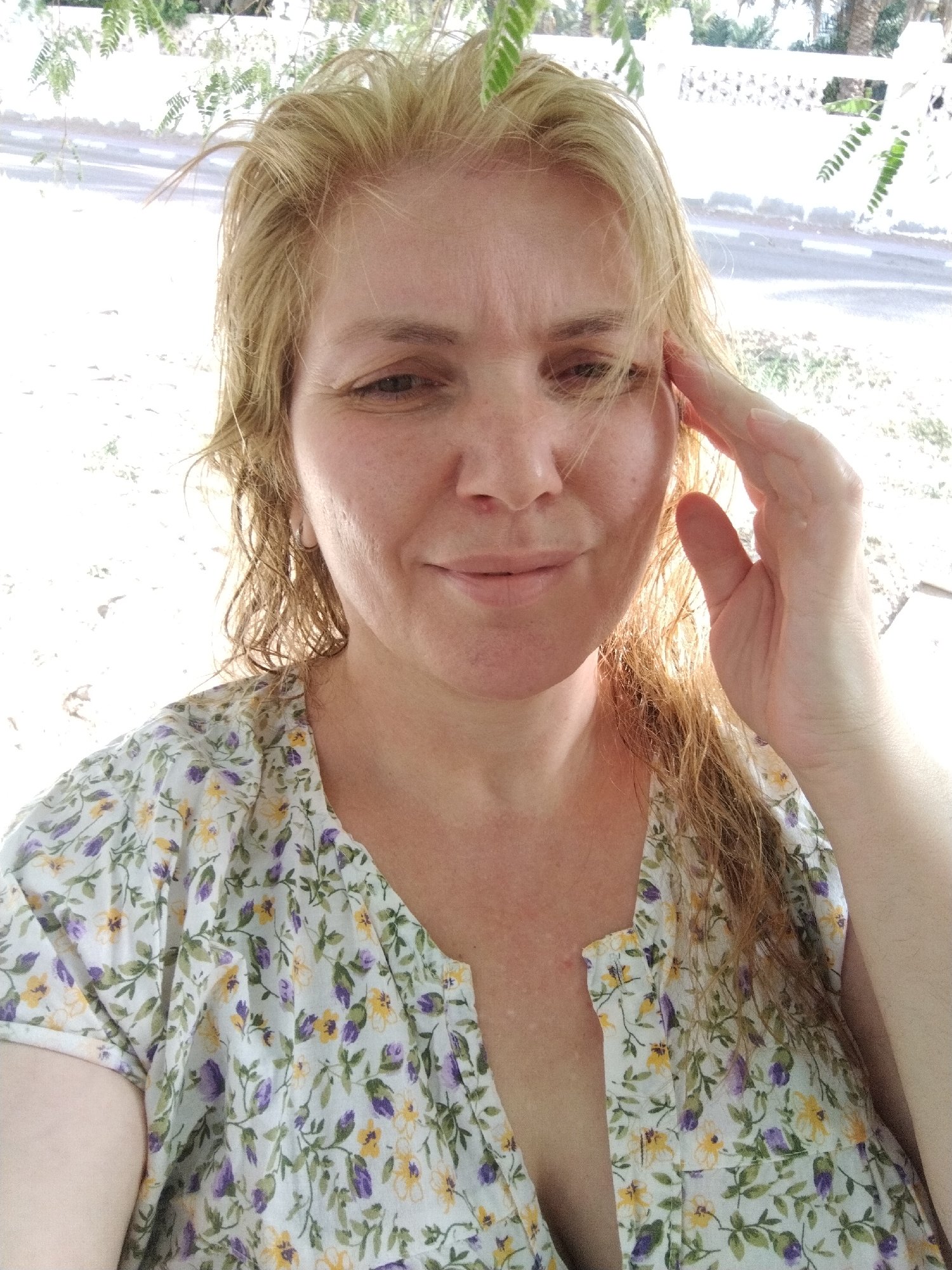 A woman with blonde hair poses for a selfie in Al Mas'oudi, United Arab Emirates. She is wearing a floral-patterned blouse, and has her hand raised near her temple. The background includes glimpses of foliage, buildings, and a street. The lighting suggests an outdoor environment.
