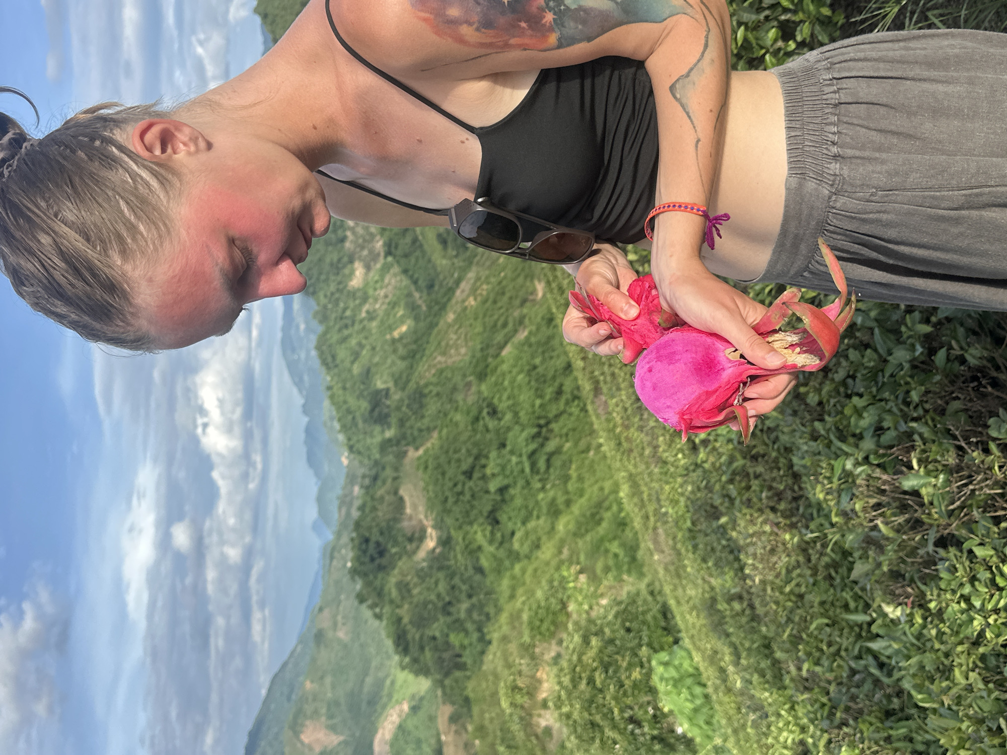 A woman stands in a lush, mountainous landscape with green terraced hills and a cloudy sky. She is wearing a black tank top and grey pants. She holds two bright pink dragon fruits in her hands, appearing to peel one. Her right arm displays a colorful tattoo. Sunglasses are tucked into the neckline of her top. A braided bracelet adorns her left wrist. The scene is captured outdoors in what appears to be a rural or natural setting.