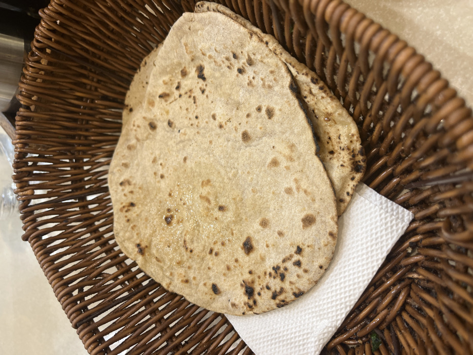 The image shows two round, flat, brown, unleavened breads called chapatis, stacked on top of each other. The chapatis are sitting on a white paper napkin in a brown wicker basket. The basket is made of woven strips of brown wood and is sitting on a plain, off-white surface. The coordinates provided correspond to the location of Sahibzada Ajit Singh Nagar, India. The image likely captures a typical scene from a local restaurant or home in Sahibzada Ajit Singh Nagar. The lighting is bright and the image appears to have been taken indoors. The image shows a simple and everyday scene, with no people or any other objects present besides the food, the basket, and the napkin. The focus of the image is on the chapatis, which appear to be fresh and ready to eat.