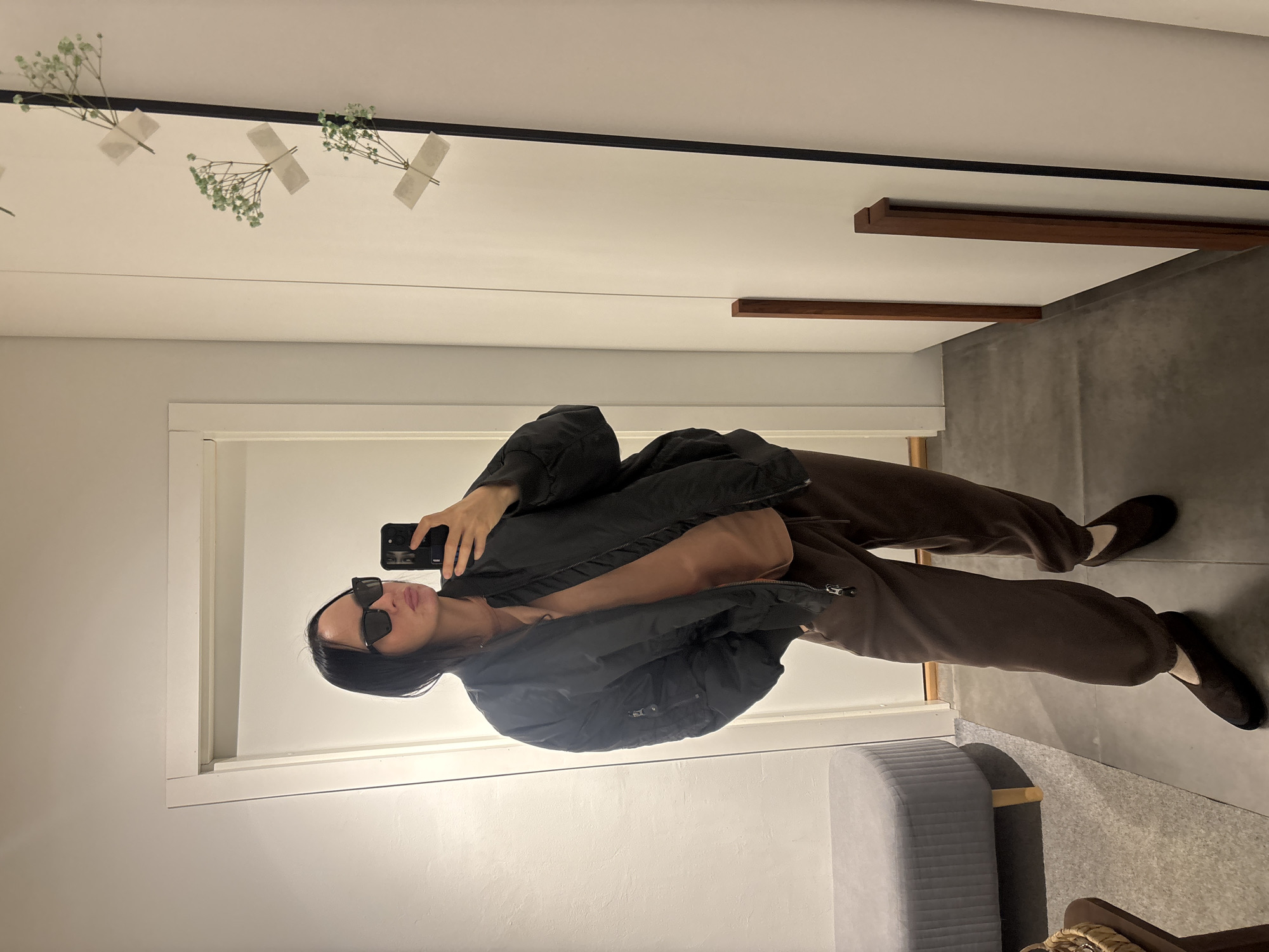 A person is taking a selfie in a room. The person is wearing sunglasses, a black bomber jacket, brown pants, and dark shoes. The room appears to be an interior space, possibly a fitting room or closet. There is a mirror reflecting the person and the room. The walls are painted white. There is a grey padded bench near the bottom of the frame. Above, on the top of the mirror frame are dried flowers attached with tape. There is no indication of Chaikovsky, Russia.