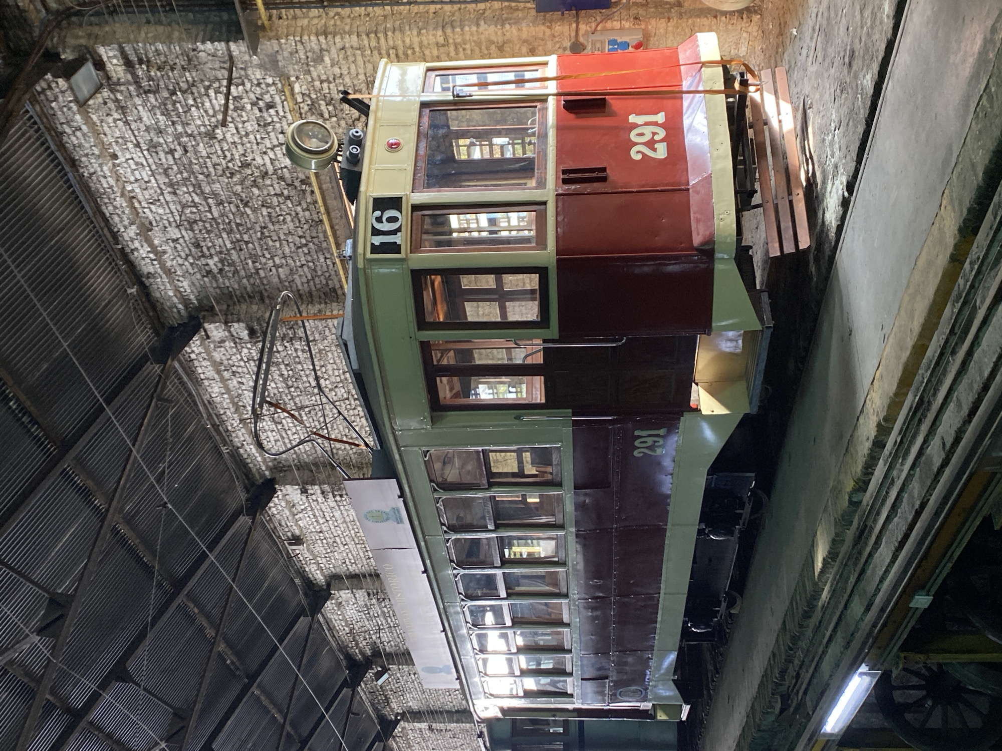 The image shows a historic tram, numbered 291 with a smaller "16" on the front, housed inside a brick and metal-roofed building, possibly a maintenance or storage facility. The tram is red and cream with multiple windows along its length. There are wooden benches on the side of the tram. The building has a weathered appearance, with exposed brickwork and a metal roof. The location is likely in the city of Buenos Aires, Argentina.