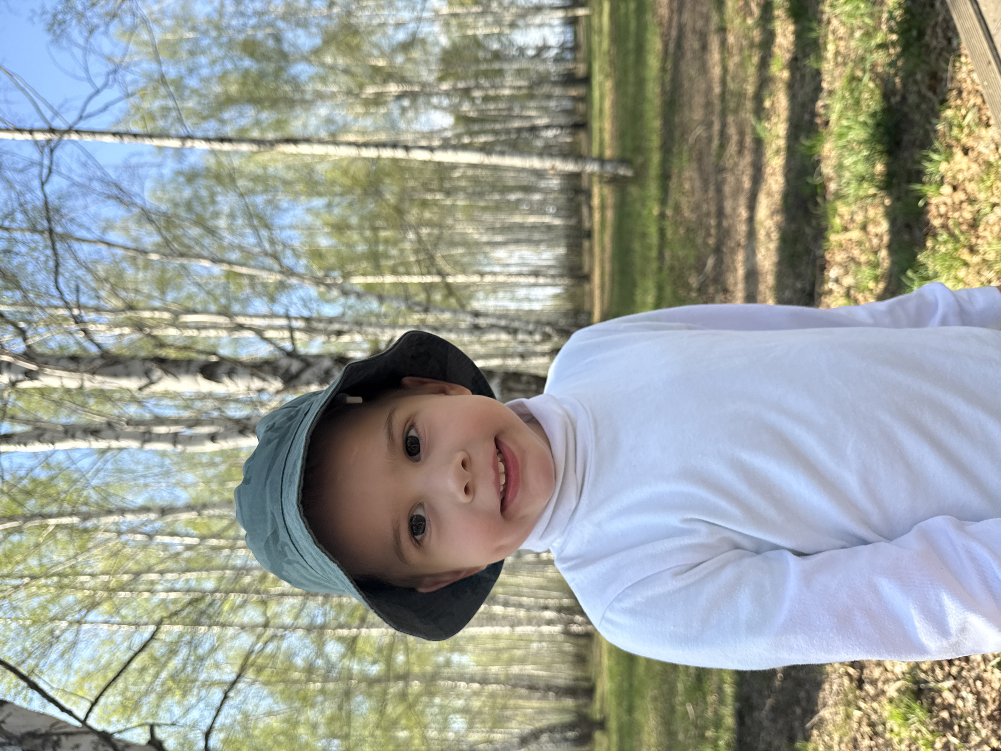 A young child wearing a white long-sleeve shirt and a green bucket hat is positioned in front of a background of birch trees. The scene appears to be outdoors in a wooded area in Chaikovsky, Russia, given the presence of the birch trees. The ground is covered with foliage and some grass. The child is looking at the camera and smiling slightly.
