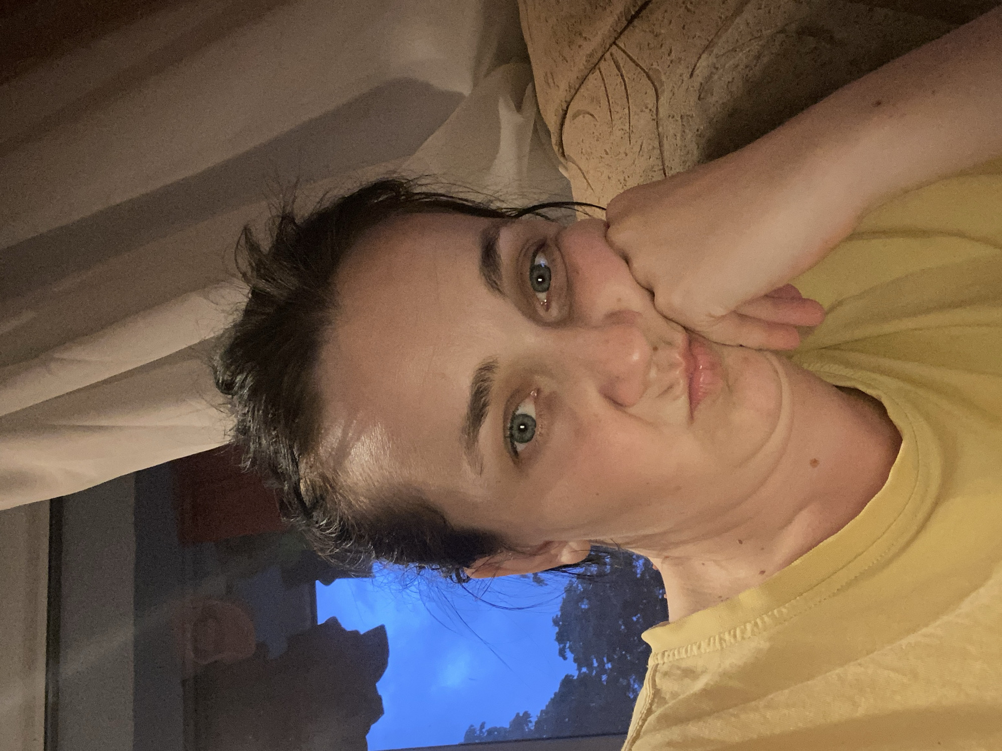 A woman with her head tilted and resting on her hand is pictured in an indoor setting. She is wearing a yellow t-shirt. Behind her, a curtain is visible, and a window shows a dark blue sky with some trees outside. The lighting is dim.