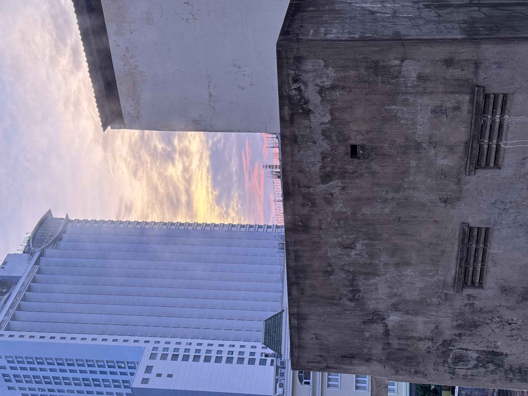The image captures a street-level view looking upwards towards a modern high-rise building with a blue facade and numerous windows, juxtaposed against an older, textured concrete building in the foreground. The sky above is illuminated by a soft sunset glow, with clouds tinged in shades of pink and orange, suggesting it is either sunrise or sunset. The concrete building in the foreground is weathered and shows signs of age, with a small rectangular opening and two rectangular vents visible on its surface. The overall scene is urban, with hints of construction visible in the background, including rebar protruding from a structure. This composition, with its mix of old and new architecture against a natural sky, is characteristic of many urban landscapes in Nha Trang, Vietnam, where rapid development often occurs alongside older structures.