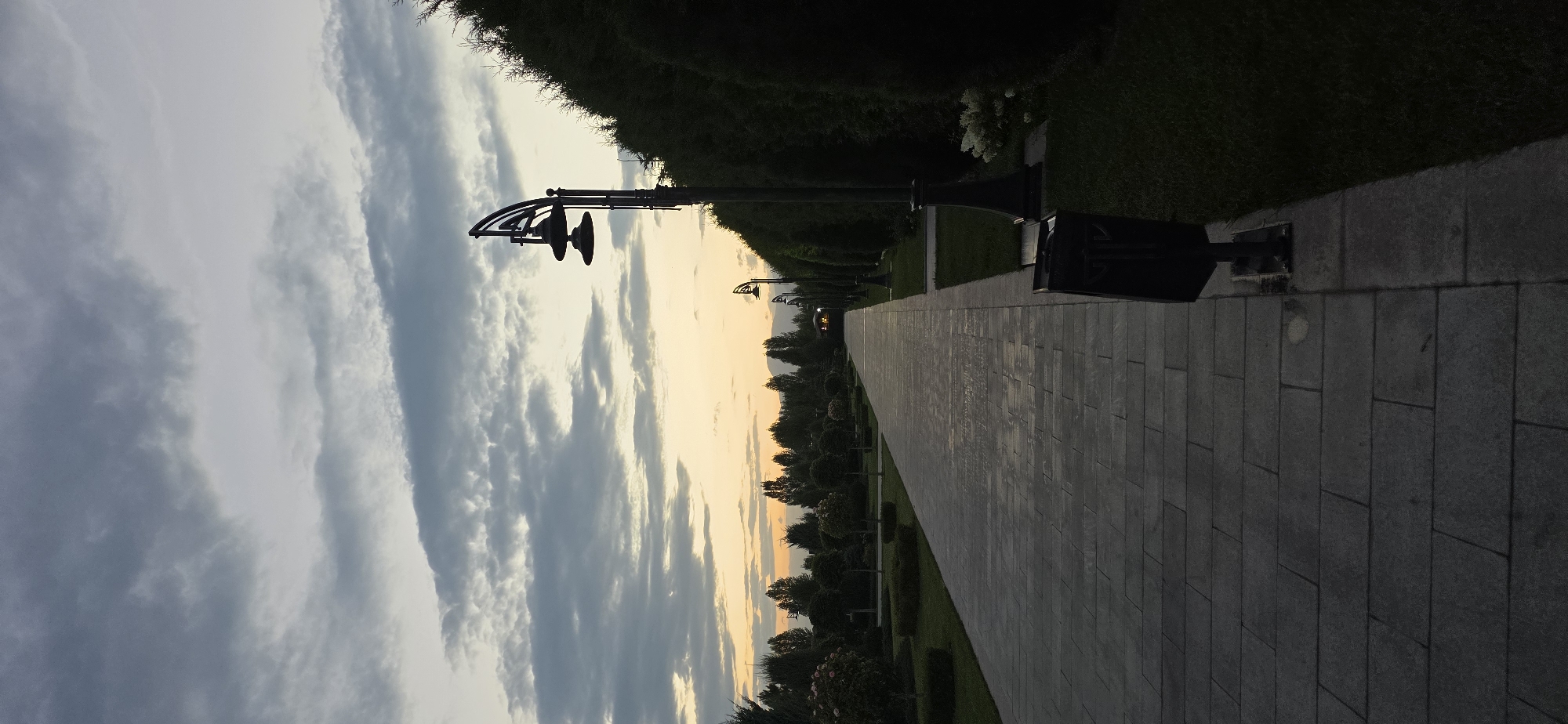 The photograph depicts a wide, paved walkway in a park-like setting, likely in Cholpon-Ata, Kyrgyzstan, during dusk. The walkway, constructed of large, rectangular stone tiles, stretches into the distance and is flanked by manicured green lawns and lush, dark green trees and bushes. On the right side of the walkway, a tall, ornate lamppost with a dark, curved design is visible, casting a faint silhouette against the twilight sky. Further down the walkway, other similar lampposts are spaced out, and a dark trash receptacle is positioned near the foreground lamppost.
The sky above is a canvas of muted blues and grays, with elongated clouds streaked across it. Towards the horizon, a soft band of pale orange and yellow light suggests the last remnants of sunset. In the background, silhouetted trees form a dense line, with the faint outline of mountains visible beyond them. The overall atmosphere is tranquil and serene, indicative of a public park or garden at the end of the day. There are no people visible in the frame.
