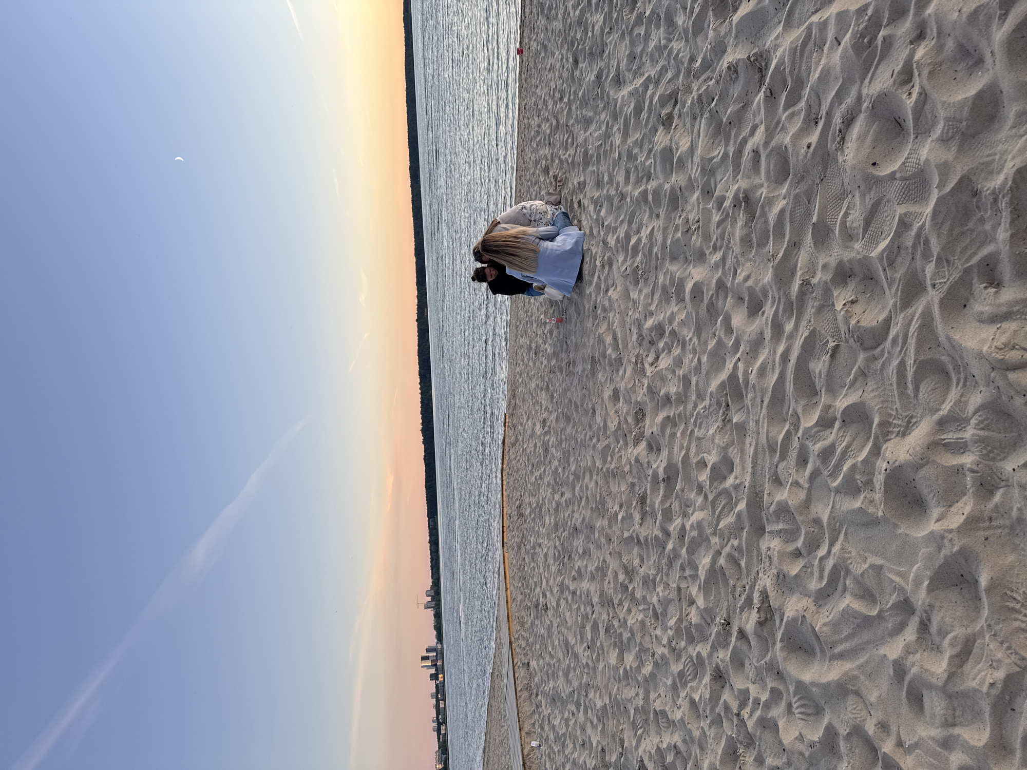 The image depicts a sandy beach next to a body of water, likely the Tallinn Bay, with the city skyline visible in the distance. The sky displays a gradient of colors from pale blue to soft orange, indicating either sunrise or sunset. A thin, wispy cloud stretches across the sky, and a crescent moon is visible in the upper right quadrant. In the foreground, the sand is textured with footprints and ripples. Three individuals are gathered on the sand, viewed from behind, appearing to be interacting. One person, wearing a light blue jacket, is kneeling. The other two are partially obscured, one in a black top and the other in a patterned garment. A pink bottle is on the sand near them. A wooden boardwalk structure is partially visible on the left side of the image, leading towards the water. The environment suggests a public beach area in Tallinn, Estonia, likely during twilight hours.