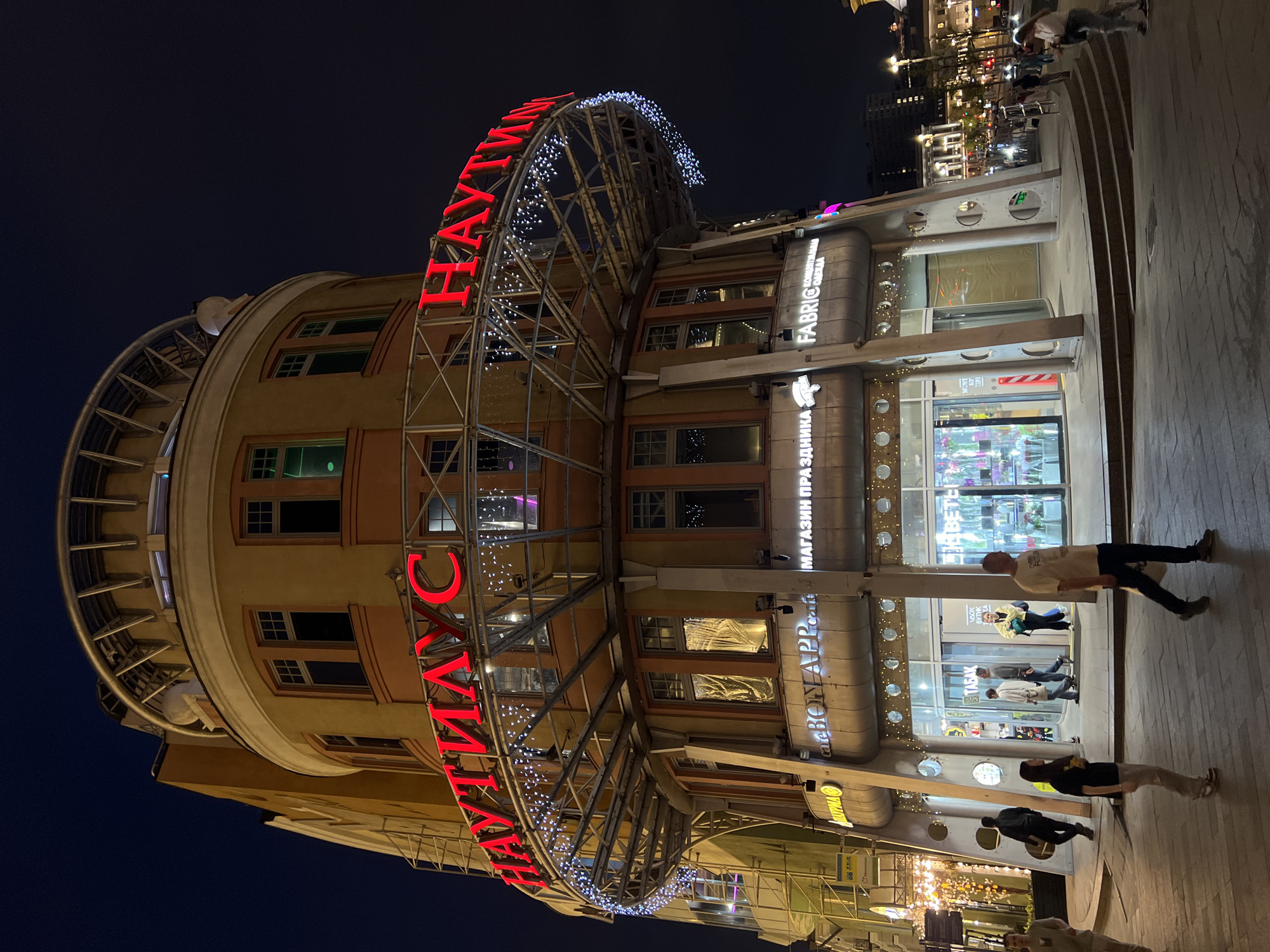 The image captures a nighttime scene in Moscow, Russia, focusing on a building with a distinctive semi-circular structure extending from its front facade. Large red Cyrillic letters, spelling out "Nautilus", arch over the entrance, adorned with strands of small white lights. The building has a beige and orange color scheme with multiple windows. Various store signs are visible on the lower floors. People are present, including a man walking in front of the building, and others further in the background. The setting is an urban environment with street lighting adding to the illumination. The ground appears to be paved. The sky is dark, indicating it is night.