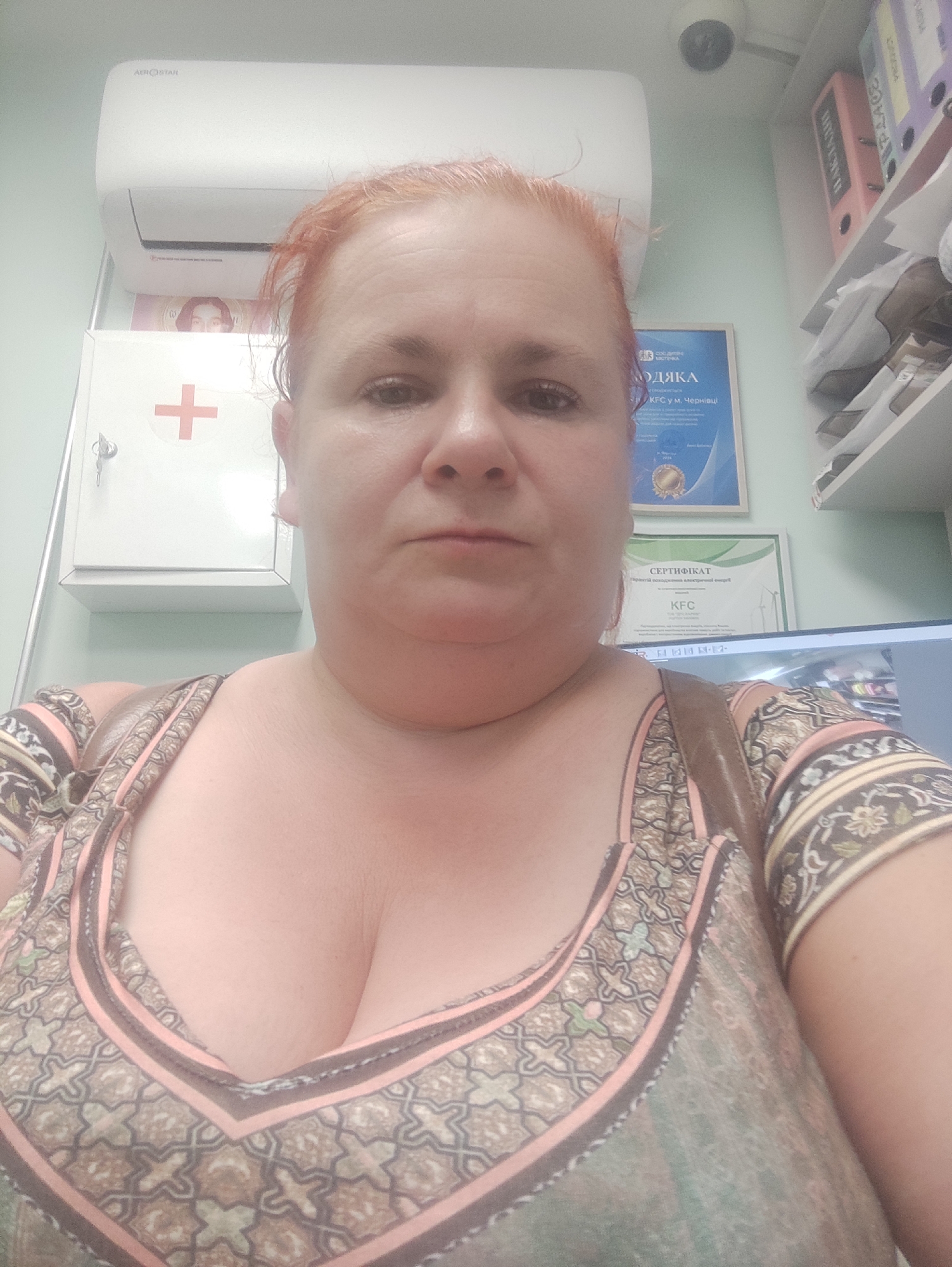 A medium shot captures a middle-aged Caucasian woman with reddish hair and fair skin, facing the camera directly. She has full cheeks and a prominent double chin. She is wearing a low-cut, patterned top with shades of pink, brown, and green. The background suggests an indoor setting, possibly an office or medical facility in Chernivtsi, Ukraine. An air conditioner is mounted high on the wall, and a white medical cabinet with a red cross emblem is visible to her left. A security camera is mounted near the ceiling. Several folders and documents are on shelves in the upper right corner, and a framed certificate with Cyrillic text is on the wall behind her. A computer monitor is partially visible in the lower right. The lighting appears to be overhead and somewhat harsh.