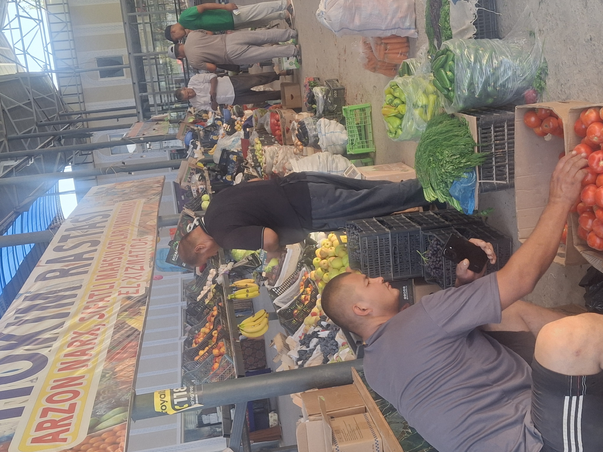 The image depicts an outdoor market stall, likely in Ko`kdala, Uzbekistan, filled with a variety of fresh produce. A man wearing a grey t-shirt and shorts is crouching in the foreground, reaching into a cardboard box filled with ripe red tomatoes. To his right, a black plastic crate contains more of these tomatoes. Behind him, a large pile of dill or fennel is displayed on a black crate, next to a clear plastic bag filled with green bell peppers and another bag containing cucumbers. In the background, other market-goers are visible browsing the stalls. Some men are seen standing near displays of fruits and vegetables, including bananas, apples, and onions. Numerous crates and bags of produce are stacked throughout the scene, indicating a busy marketplace. A large banner with text in a foreign language is displayed above the stalls. The overall environment suggests a typical agricultural market setting.