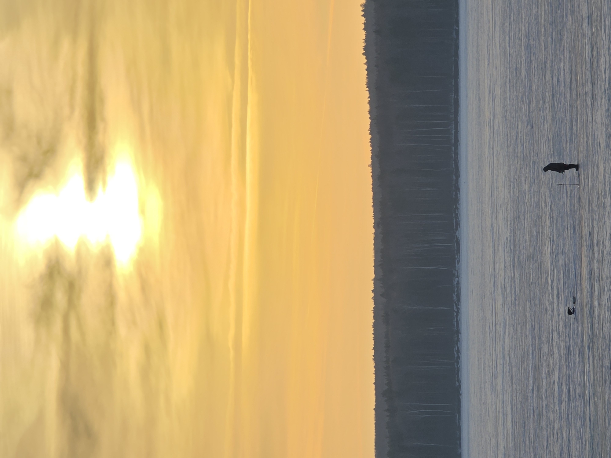 The image depicts a twilight or sunset scene at the Kama Reservoir in Chaikovsky, Russia. The left side of the frame is dominated by a bright, hazy sky with shades of yellow and orange, indicating the presence of the sun. A dark, forested shoreline is visible in the middle, separating the sky from the reservoir. The water surface is relatively calm, with a hint of gray color. On the right side, a person is standing or wading in the shallow water near what appears to be a fishing rod. There is another, smaller, indistinct object near the person in the water.