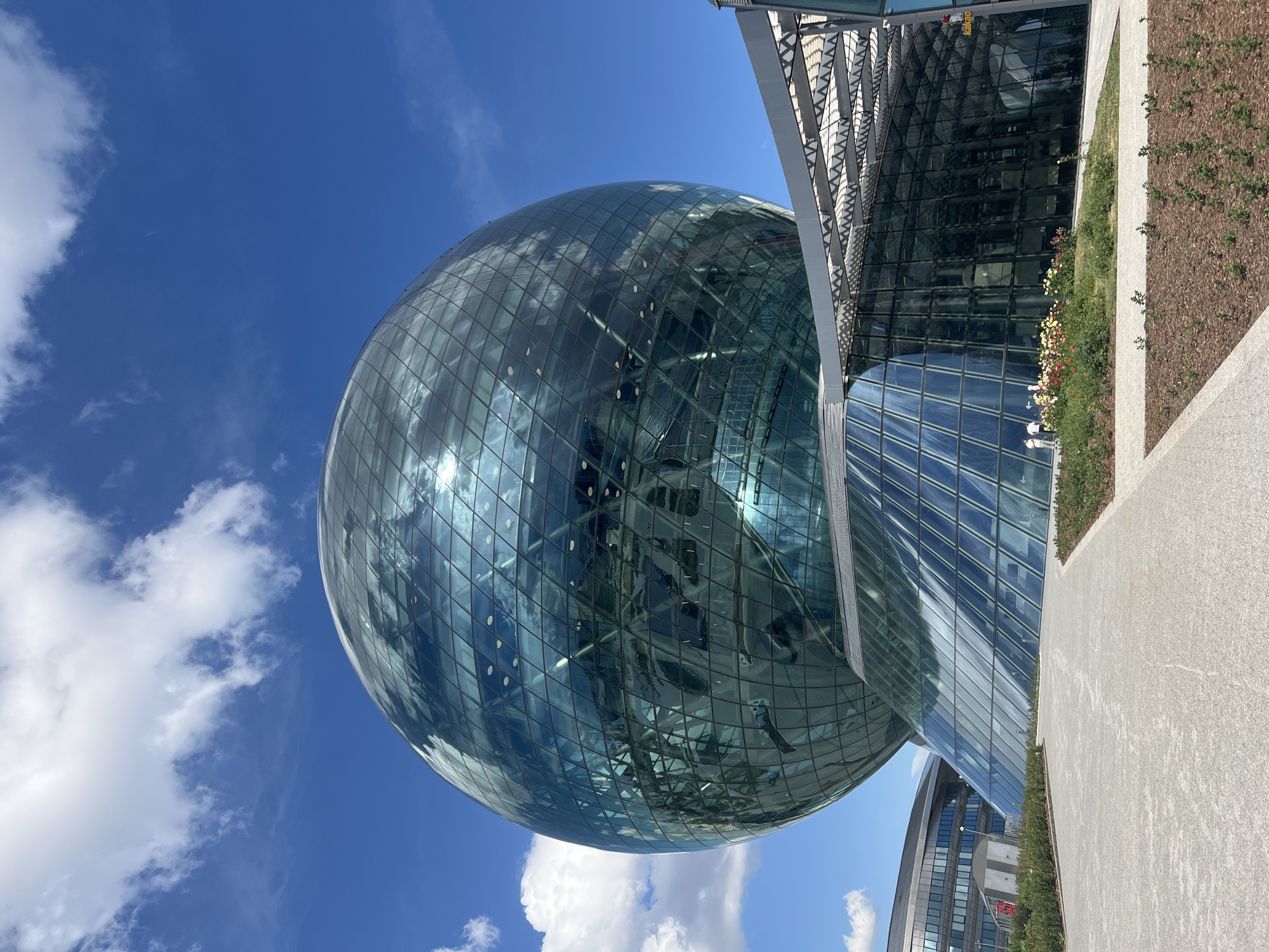 A low-angle, medium shot captures the exterior of a spherical, glass-encased building under a bright blue sky with scattered white clouds. The building, which resembles a giant glass orb, is multifaceted, with a grid-like pattern of glass panels forming its surface. Reflections of the sky and clouds are visible on the glass. The sphere is supported by a sloped, glass-covered structure that connects to a larger building with a dark, reflective facade. In the foreground, a paved pathway leads towards the building, bordered by a strip of landscaping featuring wood chips and low-lying green plants, with a few colorful flowers near the base of the sloped glass structure. Two individuals, appearing as small figures from this distance, stand near the flowers. The image appears to be taken in Astana, Kazakhstan, specifically of the "Nur Alem" pavilion, which is part of the EXPO 2017 site and now houses the Museum of Future Energy.
