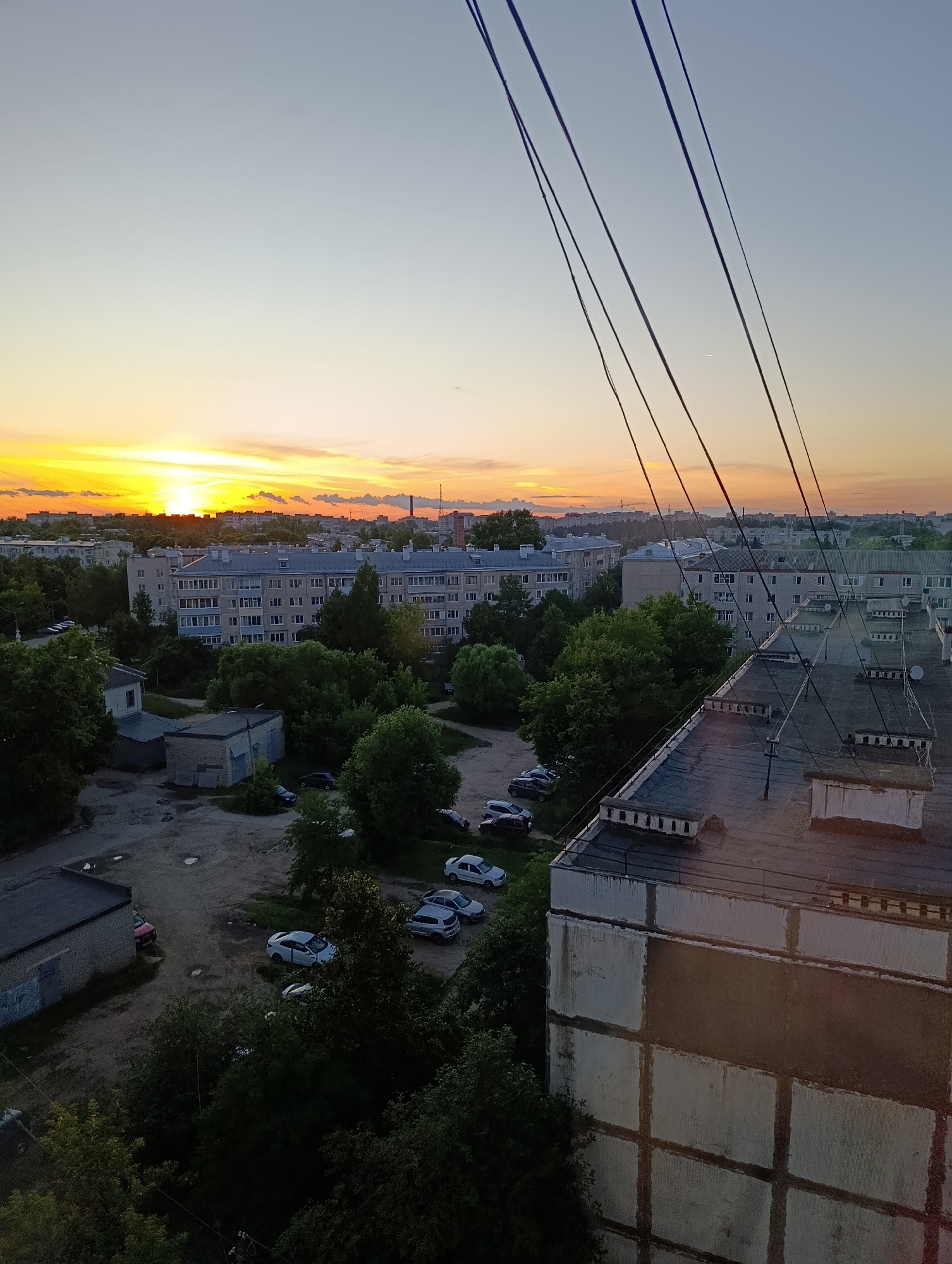 An elevated view captures a sunset over the city of Alexandrov, Russia. The sky transitions from a pale blue at the top to vibrant yellows and oranges near the horizon, with thin clouds scattered across. In the foreground, a building's rooftop is visible, featuring a dark surface with visible fixtures and a railing. Several power lines cut diagonally across the frame from the upper right to the lower left.
Below the rooftop, a dense canopy of green trees spreads across the midground. Several apartment buildings, characterized by their light-colored facades and numerous windows, are nestled among the trees and extend into the background. A parking lot is visible on the left side, containing several parked cars of varying colors, including white, silver, and dark hues. Adjacent to the parking lot are smaller, low-rise buildings, one with a grey, textured exterior and a large closed garage door. Another building on the far left has a similar grey facade. The ground in the parking area appears to be asphalt, with some puddles visible in the lower left portion of the image. The overall environment depicts a residential urban setting at dusk.
