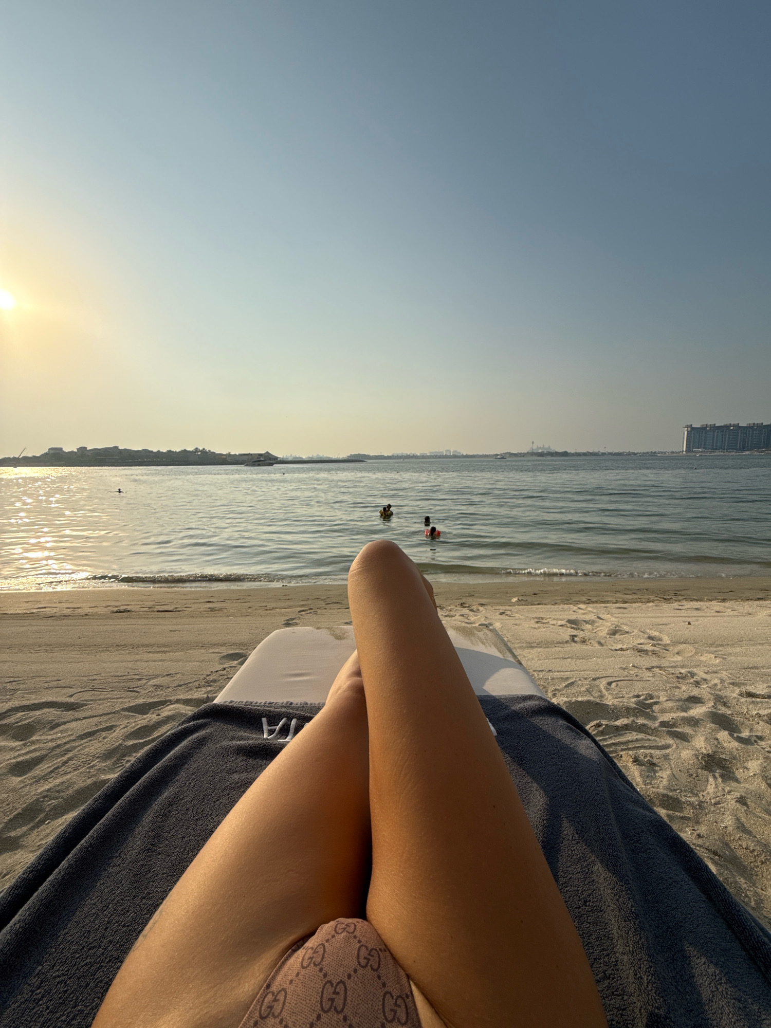 The image shows a point-of-view shot of a person lying on a beach lounger on a sandy beach. The person's legs are crossed and visible in the foreground, wearing a patterned bikini bottom. A dark gray towel is draped over the lounger. In the background, the calm ocean stretches out towards the horizon. Several people are visible in the water, some appearing to be swimming. The sun is setting, casting a warm glow on the water and sky, which transitions from a soft yellow near the horizon to a pale blue. In the distance, a skyline of buildings and structures is visible, suggesting an urban coastal environment. The overall scene depicts a relaxing day at the beach in Dubai, United Arab Emirates, likely during the late afternoon or early evening.