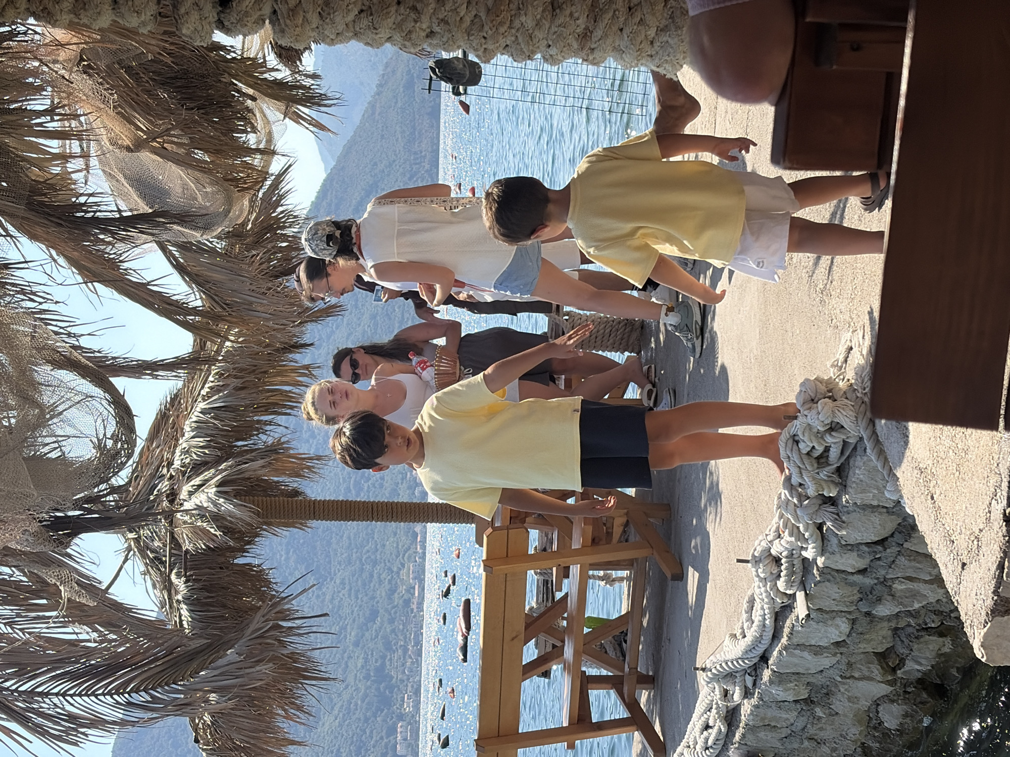 The image depicts a group of people on a sunny day, likely at a waterfront establishment in Dražin Vrt, Montenegro. Two young boys in yellow t-shirts are prominently featured in the foreground. The boy on the left wears dark shorts, while the boy on the right wears white shorts. Behind them, several adults are standing and conversing. One woman in a white top and denim shorts is visible, and another woman in a white tank top is holding a basket. The background shows a body of water with buoys, and beyond that, lush green hills under a clear blue sky. Palm trees with dried fronds provide shade, and a wooden table and benches are set up near the water. The scene suggests a relaxed, outdoor gathering by the sea.