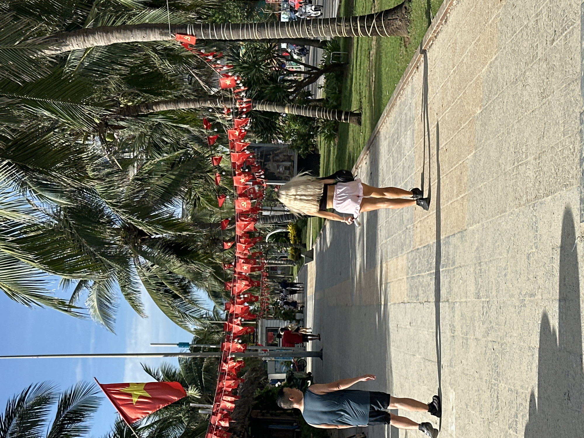 The image depicts a sunny day on a paved walkway lined with palm trees and decorated with red triangular flags. The walkway is in Đà Nẵng, Vietnam, as indicated by the Vietnamese flag visible on a flagpole. A man in a dark tank top and shorts walks away from the camera on the left side of the walkway. Further down the path, a woman with long blonde hair, wearing a black crop top and pink ruffled shorts, also walks away from the camera. Several other people are visible in the distance, walking along the path and on the adjacent street. The environment suggests a tropical tourist area, likely a beachfront promenade, with shops and greenery.