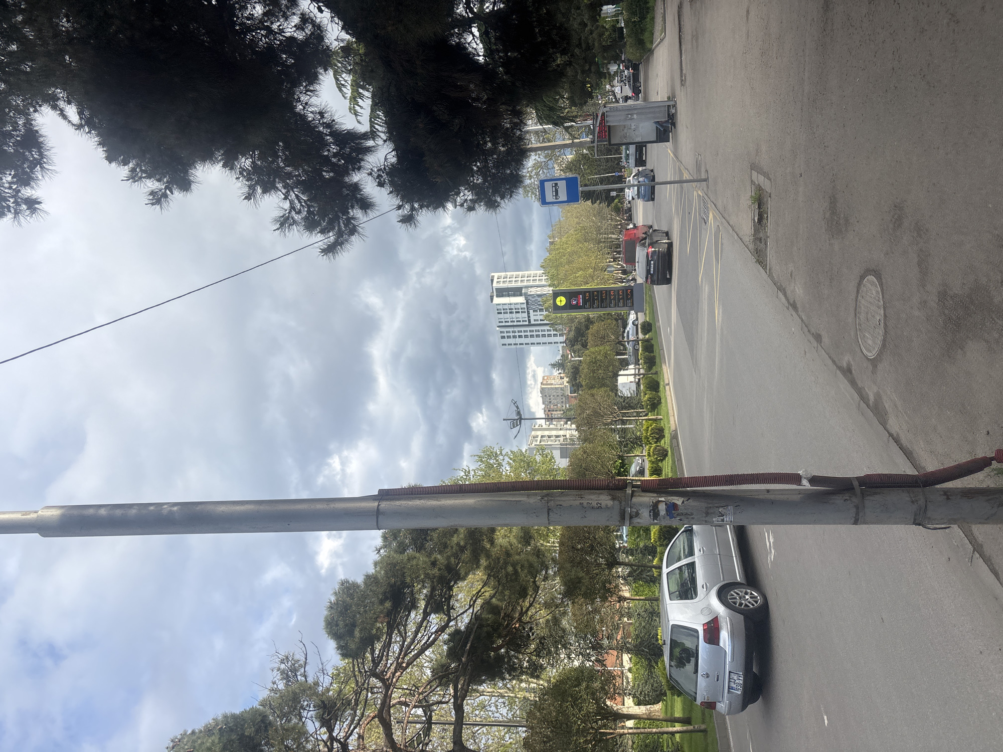 The image shows a street scene in Tbilisi, Georgia. In the foreground is a metal utility pole next to a parked silver Volkswagen Golf. Beyond the parked car, a street runs into the distance, lined with trees and buildings. A bus stop is visible along the road, along with other parked and moving vehicles. In the background, tall residential or office buildings rise against a cloudy sky. A gas station price sign is prominently displayed along the street.