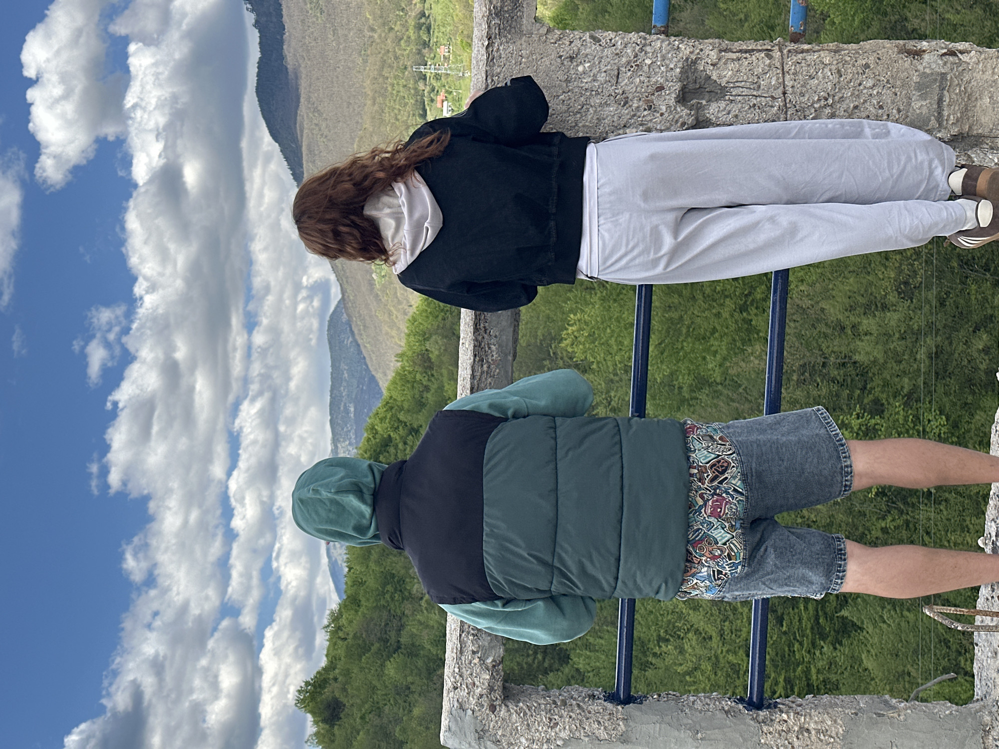 Two individuals are standing on a concrete structure overlooking a valley. The person on the left is wearing a black jacket over a white hoodie and grey pants, along with brown shoes. The person on the right is wearing a green hoodie and a vest with black and green sections, denim shorts, and tan skin. A blue railing is in front of them. The background features mountains, trees, and a cloudy sky. The location is Đurđevića Tara, Montenegro.