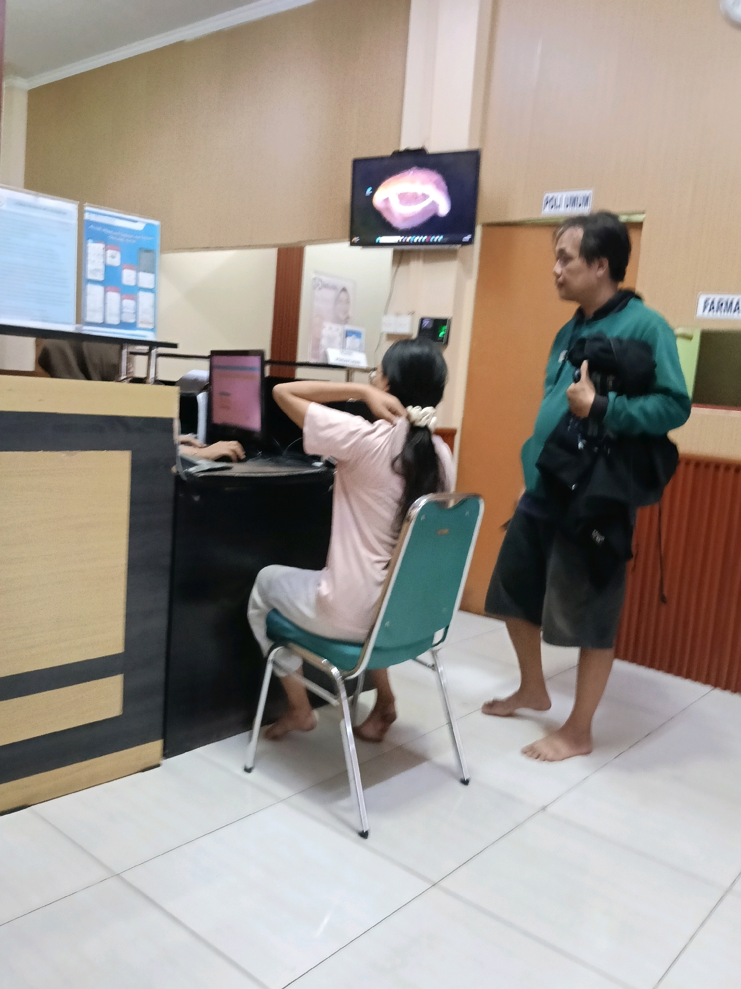 In a brightly lit indoor scene, likely a waiting area or reception of a medical facility in Kosambi Timur, Indonesia, a man and a young person are present. The man, wearing a dark green jacket over a dark shirt and dark shorts, stands on the right side of the frame, barefoot, holding a black bag in his right hand. He looks towards the left, his gaze directed at the young person. The young person, seated on a teal-colored chair facing a desk with a computer, is wearing a light pink t-shirt and white pants. They are barefoot and appear to be interacting with the computer, with their left hand on the keyboard. A black computer monitor with a pink screen is visible on the desk. In the background, a television screen displays a close-up image of teeth. Signs on the wall indicate "POLI UMUM" and "FARMA". The environment is clean with tiled flooring.