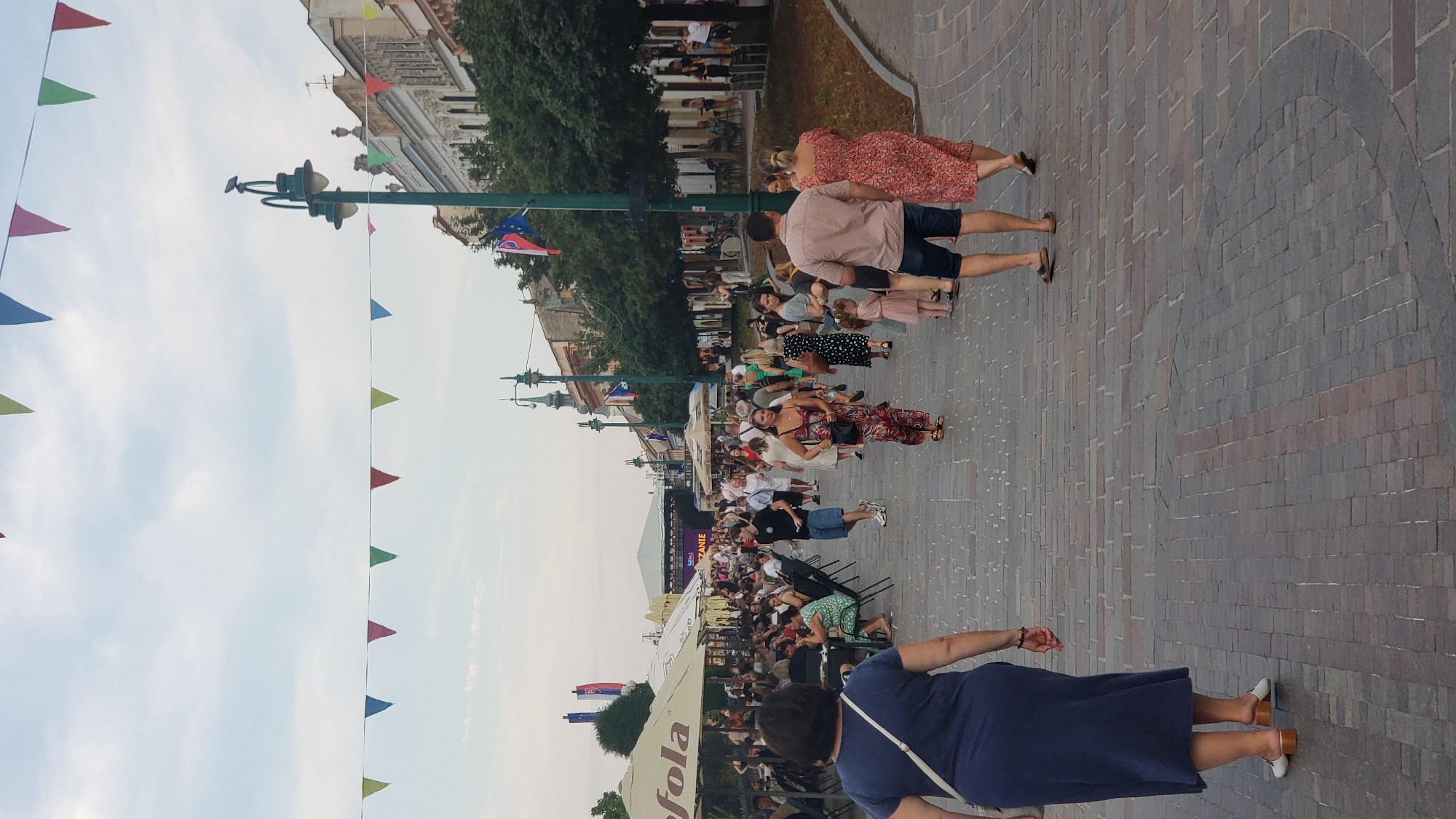 The image depicts a street scene in Prešov, Slovakia, likely during a public event or festival. Numerous people are strolling along a brick-paved pedestrian walkway lined with buildings and trees. Several stalls or outdoor dining areas with umbrellas are visible, suggesting food and beverage services. Colorful triangular flags are strung across the sky, adding to the festive atmosphere. In the background, a stage is set up, hinting at a performance or concert. Two Slovak flags are prominently displayed on a lamppost. The lighting suggests it is daytime, possibly late afternoon or early evening, given the soft light. The overall impression is one of a lively, community gathering in a European city.