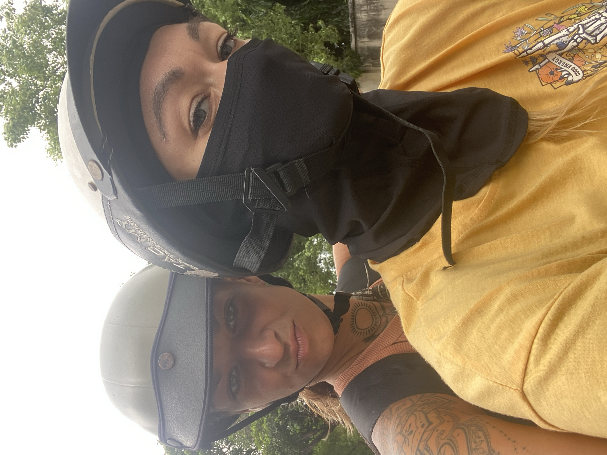 Two women wearing helmets are posing for a selfie. The woman in the foreground has a light-colored helmet with a visor, a sun tattoo on her neck, and visible tattoos on her arm and chest. The woman in the background is wearing a black helmet and a black face covering, revealing only her eyes. The background includes green trees and foliage, suggesting an outdoor location. The image was likely taken in Đồng Nai, Vietnam.