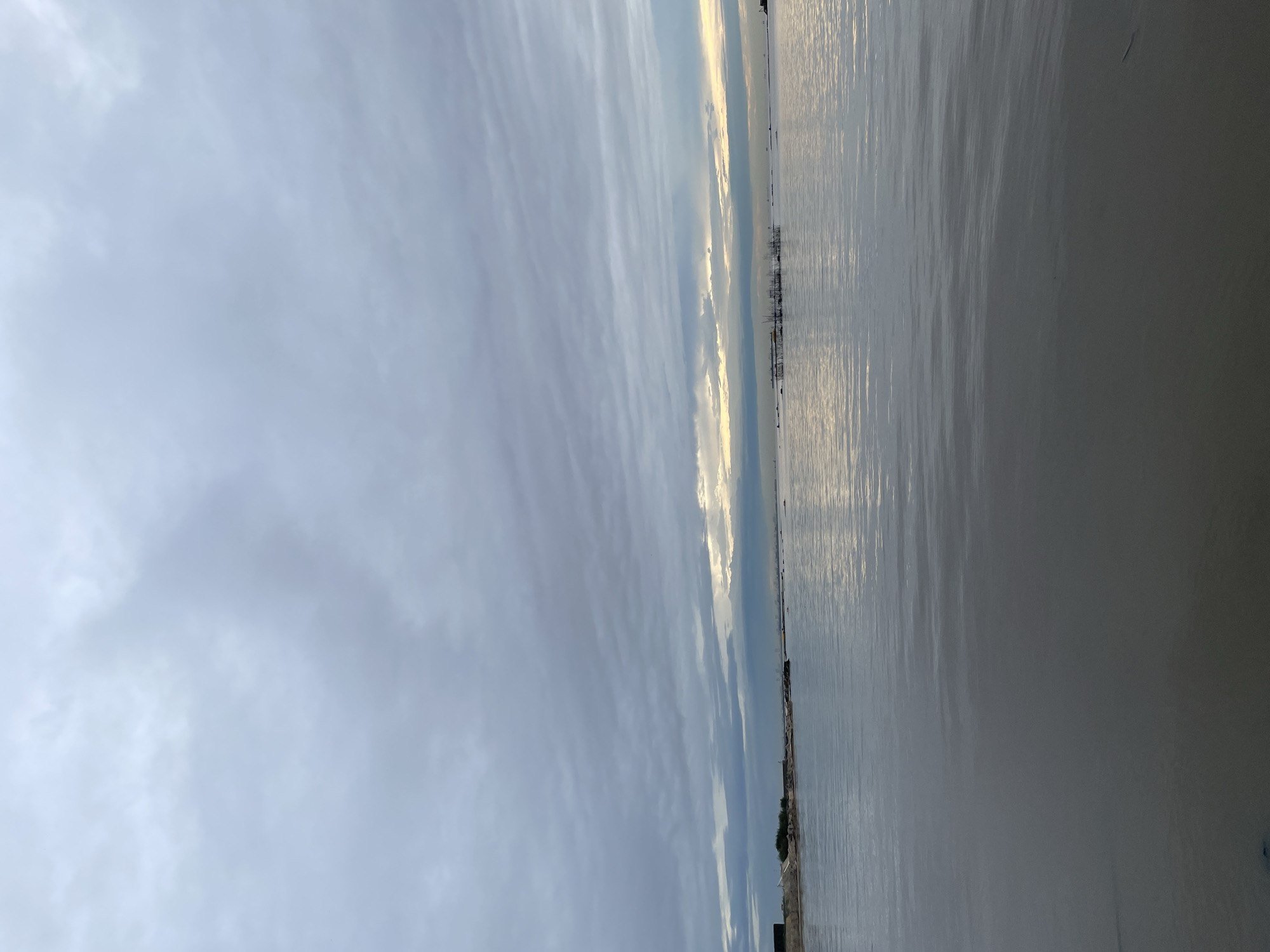 The image shows a view of the Lagos Lagoon from Ikorodu, Nigeria, during what appears to be dusk. The sky is overcast with thick, grey clouds, but there is a faint glimmer of light visible on the horizon where the sky meets the water. The water itself is calm and reflects the muted colors of the sky.
In the distance, on the left side of the frame, is a shoreline with a sandy beach and some vegetation. There are also some man-made structures, including a dark, rectangular building, and what looks like construction or debris. Further out in the lagoon, a cluster of wooden structures, possibly fish farms or floating platforms, are visible. The expanse of the lagoon stretches out to the horizon, where a faint outline of more distant land or structures can be discerned. The overall impression is one of a vast, open body of water under a moody sky, with hints of human activity along the edge and within the lagoon.
