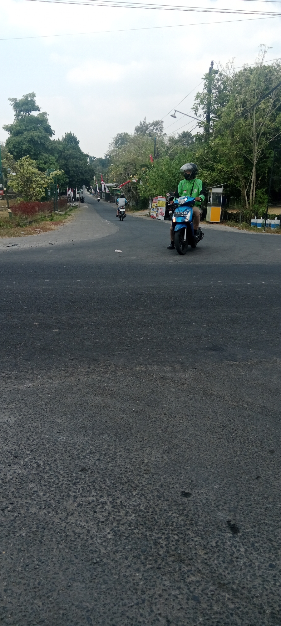 The image shows a man riding a blue motorcycle on a paved road in Maguwoharjo, Indonesia. The man is wearing a green jacket and a helmet. There are trees and flags along the side of the road. The sky is cloudy. The road appears to be a two-lane road, with one lane in each direction. There are some other vehicles visible in the distance. The overall scene suggests a rural or suburban area.