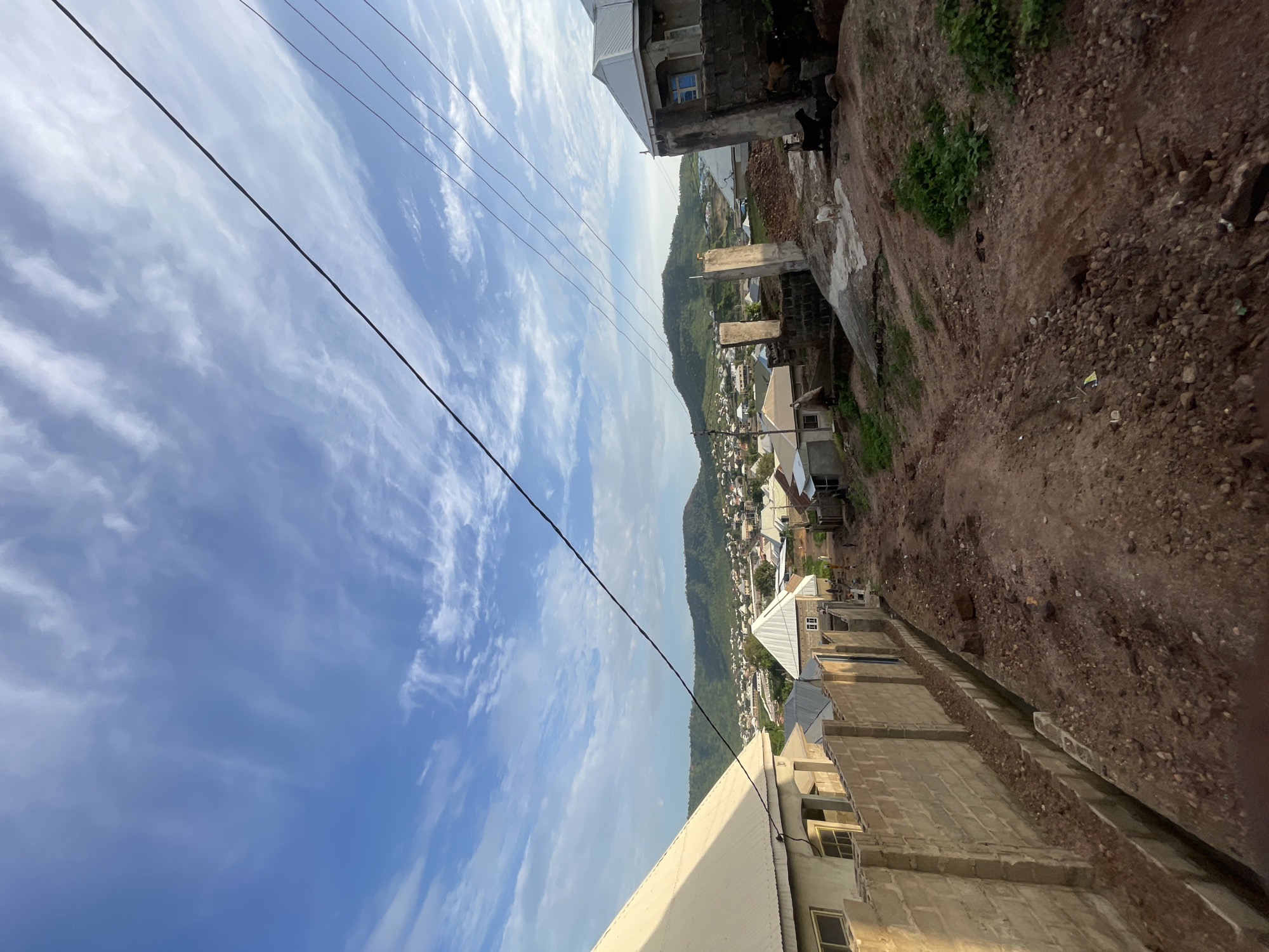 This image captures a wide-angle, elevated view of a residential area set against a backdrop of green hills, consistent with the landscape of Lokoja, Nigeria. The scene is presented from a high vantage point, looking down into a valley or settlement.
In the foreground, parts of several buildings are visible. At the bottom, a white-roofed structure indicates a finished building. Along the right side, structures made of unpainted concrete blocks suggest ongoing construction or a more utilitarian style, with some walls appearing unfinished. The ground surrounding these structures is primarily reddish-brown dirt and gravel, interspersed with small patches of green vegetation. Several domestic animals, including a black goat and a white animal (possibly another goat or sheep), are visible near one of the buildings on the upper right, situated on the unpaved terrain.
The midground reveals a denser cluster of residential buildings, extending towards the base of prominent, lush green mountains or hills that dominate the horizon. These buildings also predominantly feature concrete block construction and corrugated metal roofs.
The sky occupies the upper portion of the image, displaying a clear blue expanse with scattered white, wispy clouds. Several dark utility lines or power cables stretch across the sky from left to right, intersecting the view. The bright and even lighting indicates it is daytime, likely morning or late afternoon, suggesting fair weather. No specific human activity or visible text is present, offering a static, observational view of the urban-natural interface in this region.