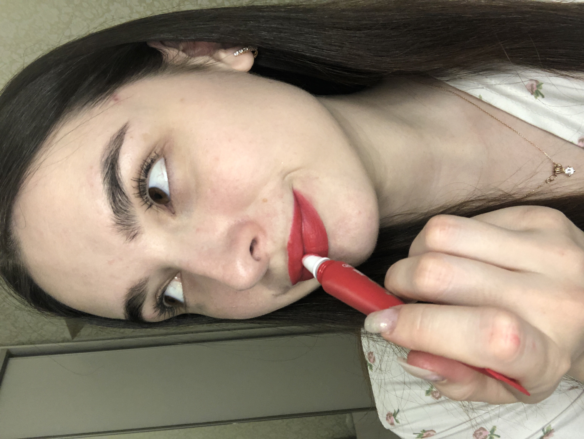 A woman with long, dark brown hair is applying red lipstick from a red tube. She has brown eyes and fair skin. She is wearing an earring on her left ear and a necklace. Her shirt has a floral pattern. A pale beige wall and a light-colored cabinet are visible in the background. There is no evidence of Noviy, Russia in this image.