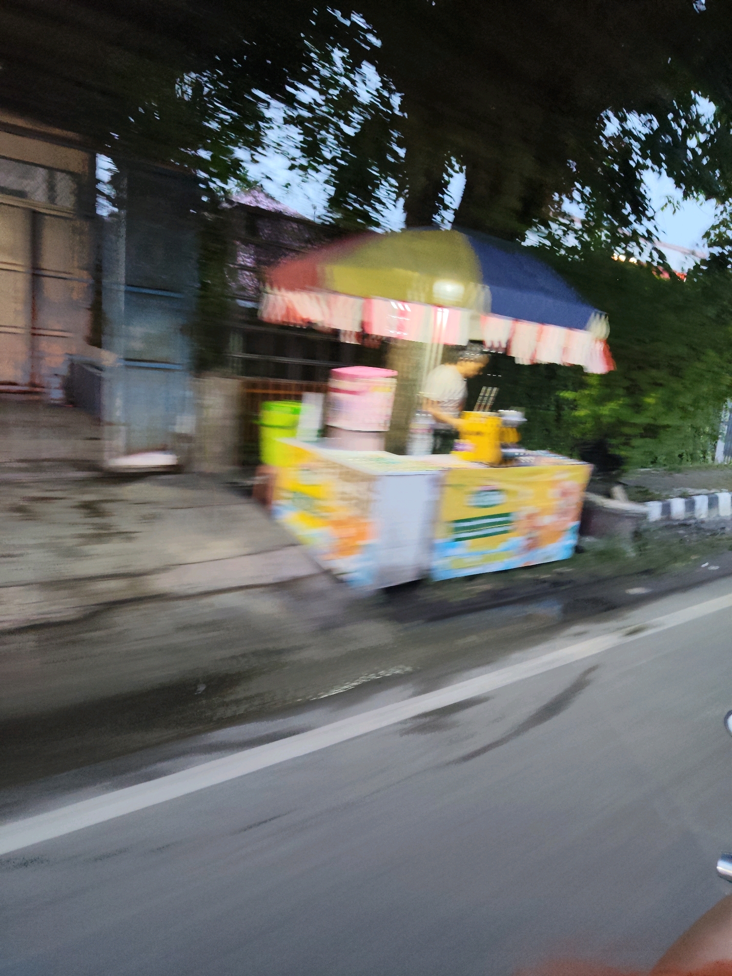 The image shows a street scene in Kaduagung Timur, Indonesia. A vendor cart with a colorful striped umbrella is visible on the side of the road. The cart appears to be selling some sort of food or drink, with several containers and a person standing behind it. There is a building to the left of the cart and trees and foliage to the right. The road is wet, suggesting recent rain. The image is slightly blurry due to motion, indicating it was likely taken from a moving vehicle.