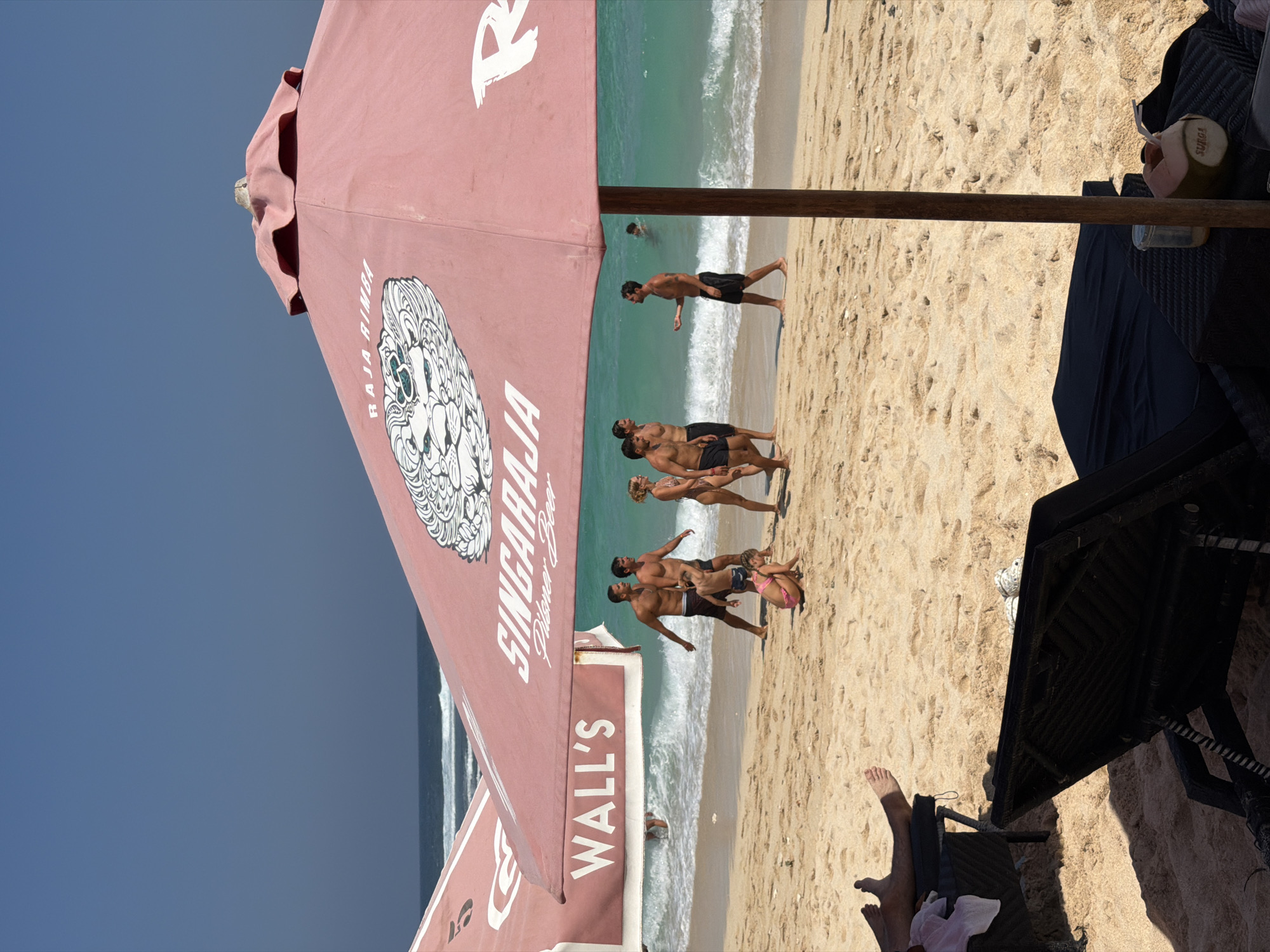 The image depicts a sunny day at a beach. In the foreground, there are two empty beach chairs with dark fabric. A pink umbrella, emblazoned with the words "SINGARAJA Pilsner Beer" and "RAJA RIMBA" along with a stylized lion's head, dominates the central part of the frame, obscuring a portion of the sky and the ocean. To the left, another umbrella is partially visible, with the word "WALL'S" written on it.
On the sand, a group of people are walking near the water's edge. Two men, one wearing black shorts and the other with dark swim trunks, are walking side-by-side, with a young boy in blue shorts in front of them. To their right, a woman with blond hair in a bikini is walking. Further to the right, a man in black shorts is speaking to another man in the water. A young girl in a pink bikini is playing in the sand closer to the foreground.
The ocean is a vibrant turquoise color with white waves. The sand is light brown and appears to be fine-grained, with numerous footprints and indentations. The sky is a clear, bright blue. On one of the beach chairs in the foreground, a young coconut with a straw is placed on a table next to a small glass jar.