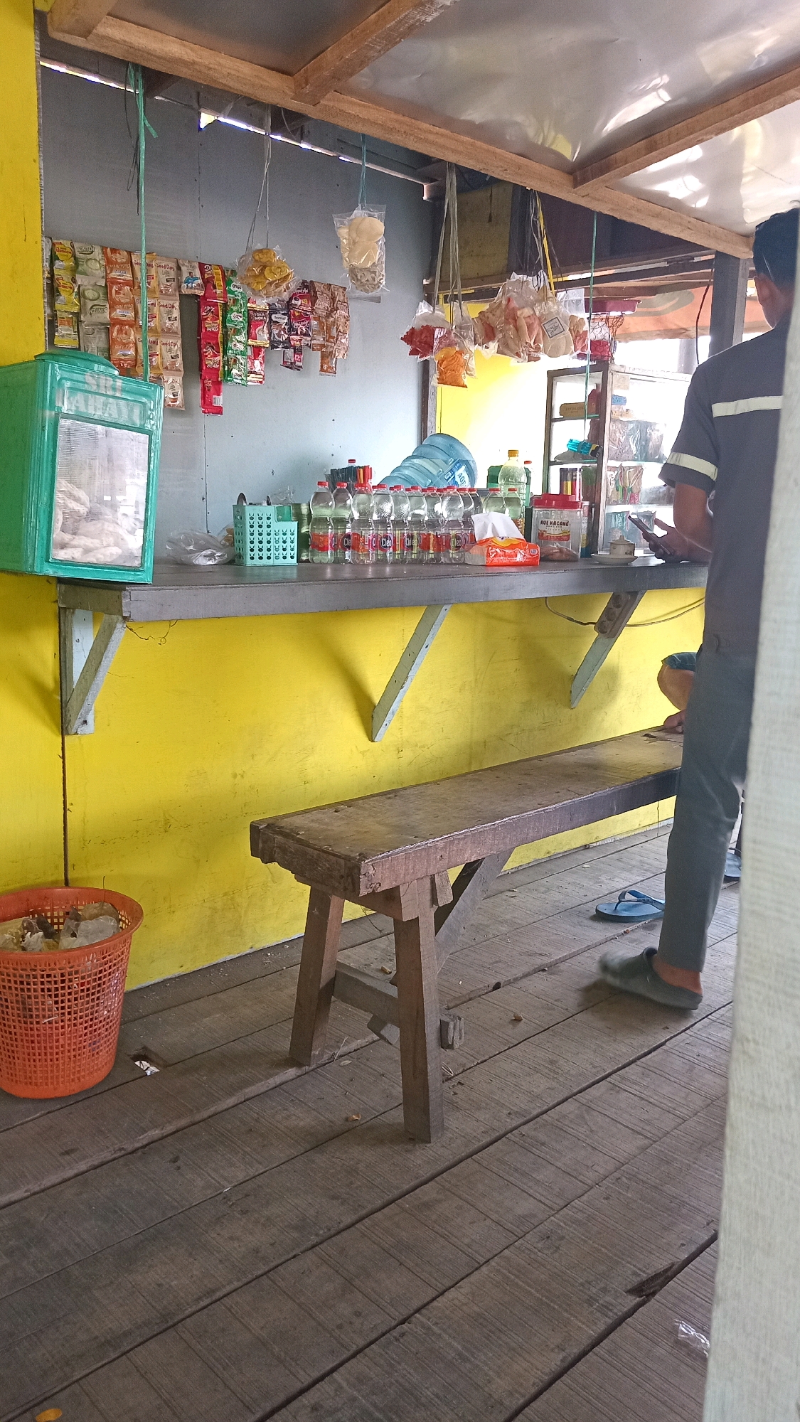 The image depicts a small outdoor kiosk located in Ambeng Ambeng Watangrejo, Indonesia. The kiosk has a bright yellow wall and a wooden counter displaying various items. Among the items on the counter are bottled drinks, a tissue box, a container of coffee or sugar, and what appears to be a calculator or phone. Several bags of snacks are hanging from the ceiling above the counter. In the foreground, a wooden bench is positioned in front of the counter, and a red plastic basket, possibly a trash can, is situated to the left. A person is partially visible on the right side of the frame, standing near the counter and appearing to be interacting with their phone. The kiosk has a rustic, open-air structure with a wooden floor.