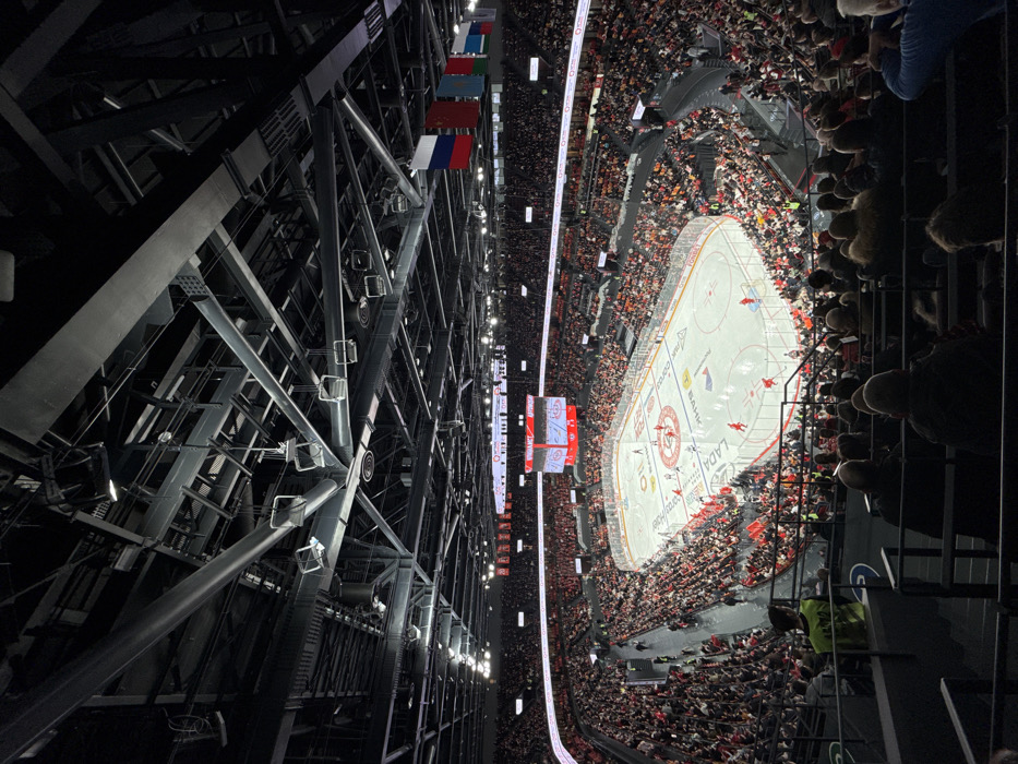 An overhead view captures an ice hockey game in progress at the Uralets Arena in Yekaterinburg, Russia. The brightly illuminated ice rink is centrally located, displaying advertising logos such as LADA, Fonbet, and Bank. Multiple players in red and blue/white jerseys are visible on the ice. A large, multi-sided scoreboard, suspended above the rink, displays game information including "АВТОМОБИЛИСТ" and "ШТОРМ" along with scores and time.
The arena seating, primarily dark, is densely populated with spectators largely facing the rink, with a notable concentration of red attire in lower sections. In the immediate foreground, the backs of several spectators' heads are visible, alongside an individual wearing a neon yellow vest on the left. High above the seating on the far side, a row of national flags is suspended, including those of Russia, China, Kazakhstan, and Belarus. The arena's ceiling structure consists of a complex network of dark steel beams, trusses, and numerous spotlights directed downwards onto the rink.