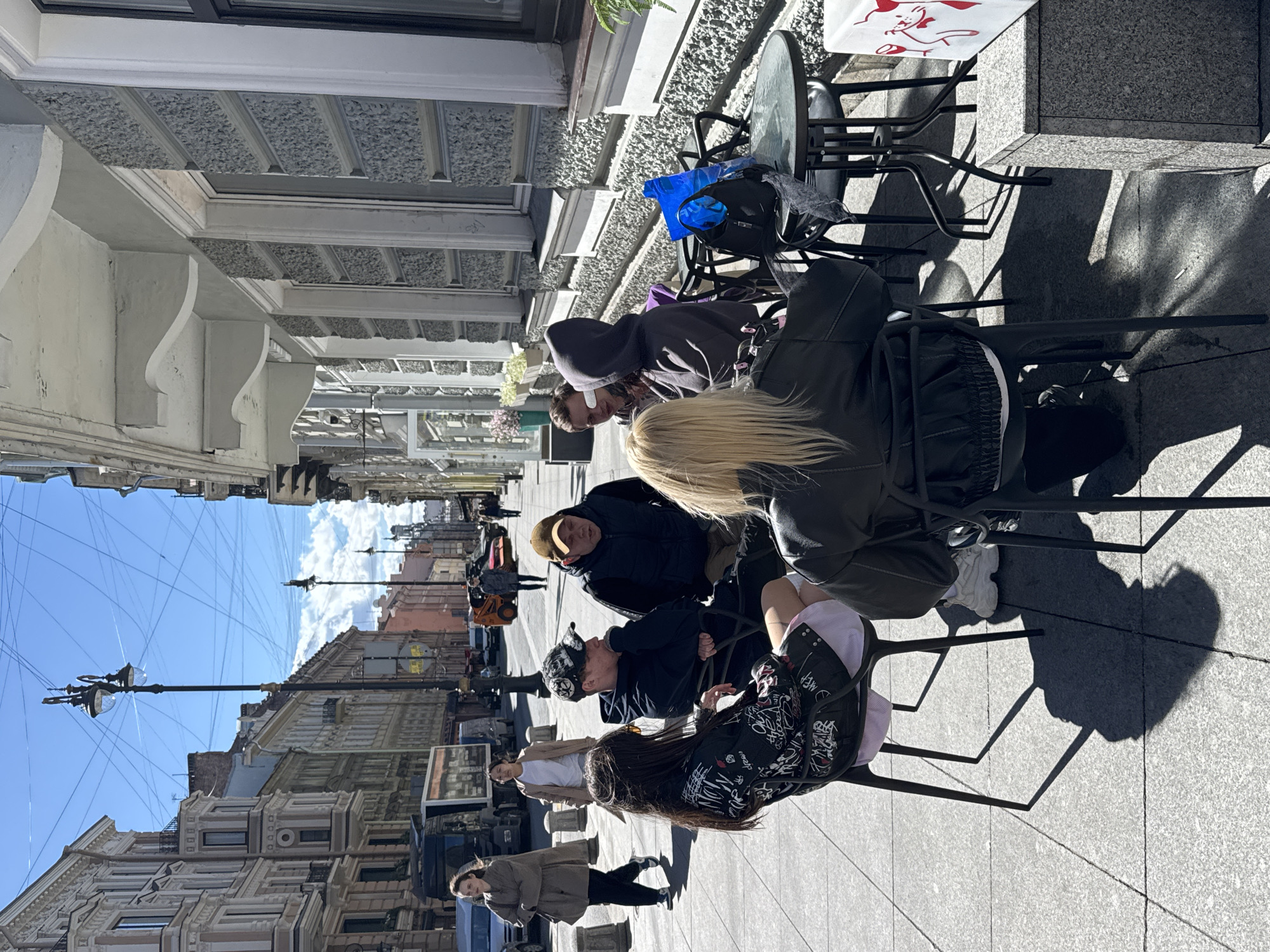 The image depicts a street scene in Saint Petersburg, Russia, on a sunny day. Several people are seated at outdoor cafe tables, while others are walking along the sidewalk. Architectural details of buildings line the street, and overhead wires stretch across the sky. At least 6 people are seated on black metal chairs around a round table. Two people are walking in the background, a female wearing a cream colored shirt and male wearing a beige jacket.