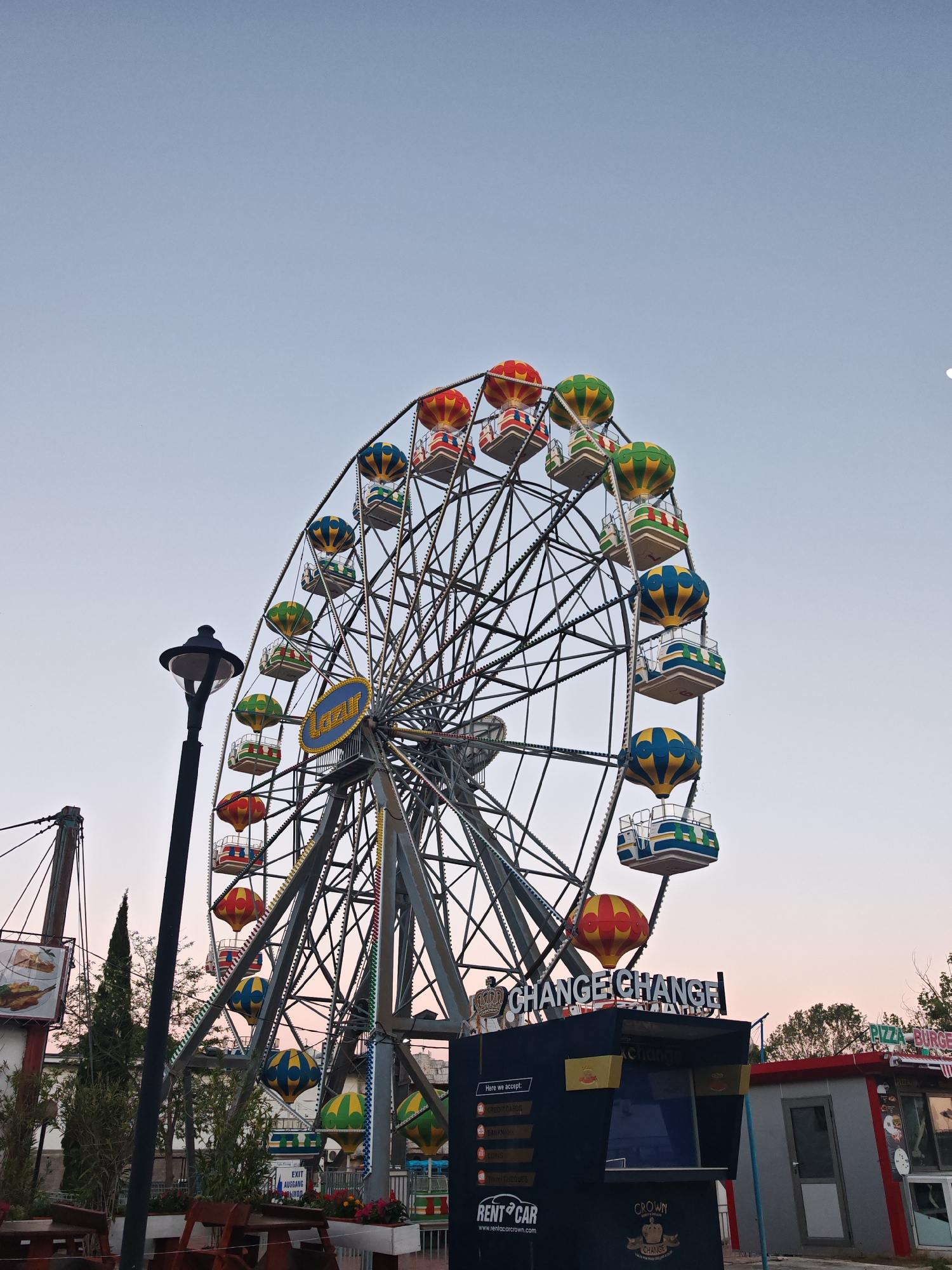 The image depicts a Ferris wheel at dusk, likely in an amusement park in the city of Nesebar, Bulgaria. The Ferris wheel has colorful, gondola-style seats resembling hot air balloons. A street lamp stands to the left of the Ferris wheel. Below the wheel are commercial kiosks, including a currency exchange ("CHANGE CHANGE") and food stands selling pizza and burgers. Signage indicates acceptance of credit cards and the availability of rent-a-car services. Trees and other buildings can be seen in the background. The moon is also visible in the upper-right corner of the image.