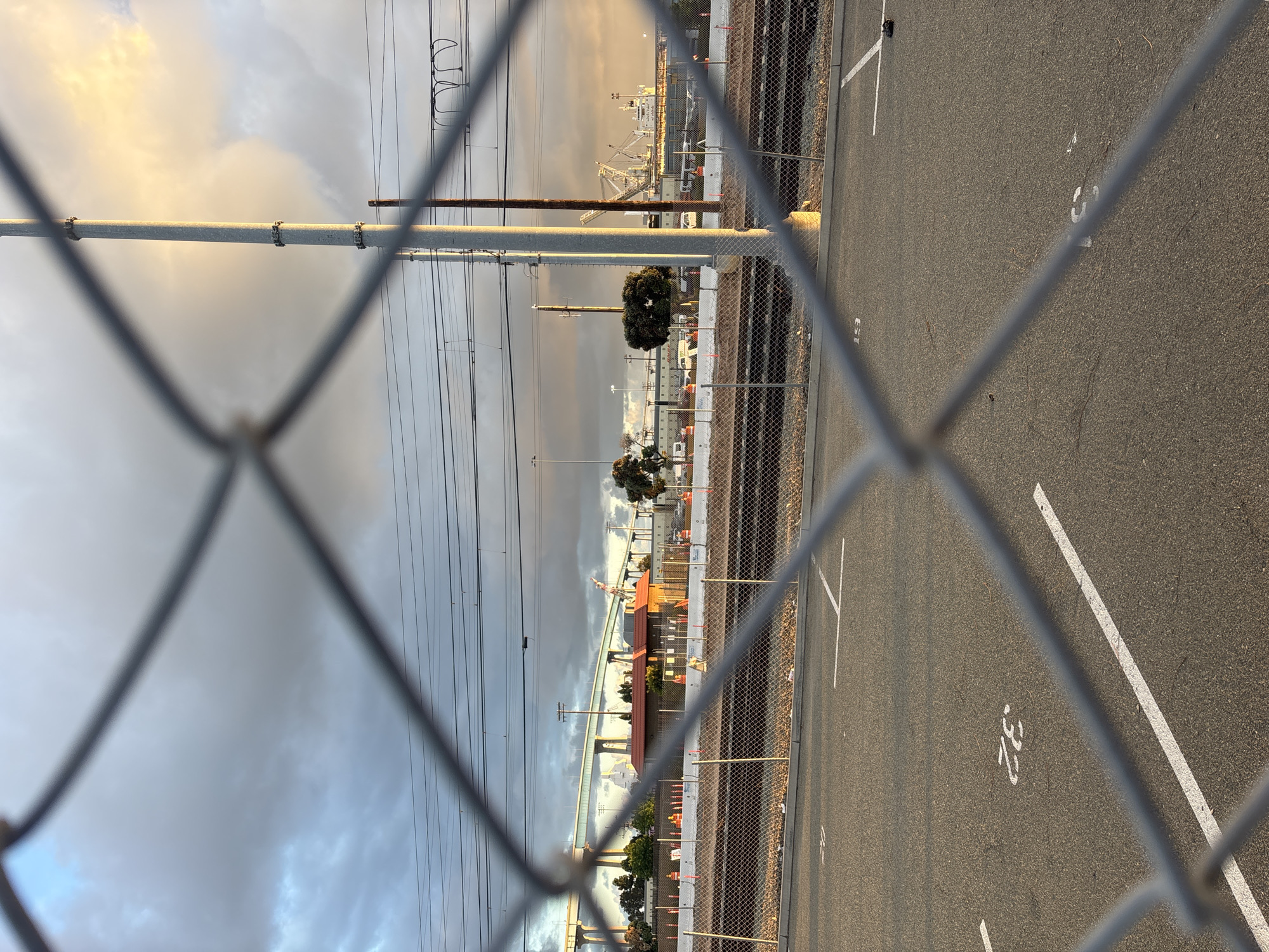 The image shows a chain-link fence in the foreground, with a parking lot visible on the right side. White lines and parking space numbers are painted on the asphalt. Beyond the parking lot is a railway track running parallel to the fence. In the distance, industrial infrastructure is visible, including cranes, buildings, and what appears to be a port facility. The sky is overcast with a mix of dark and light clouds. Power lines and utility poles are also visible in the background. The picture could be taken in the Port of San Diego area.