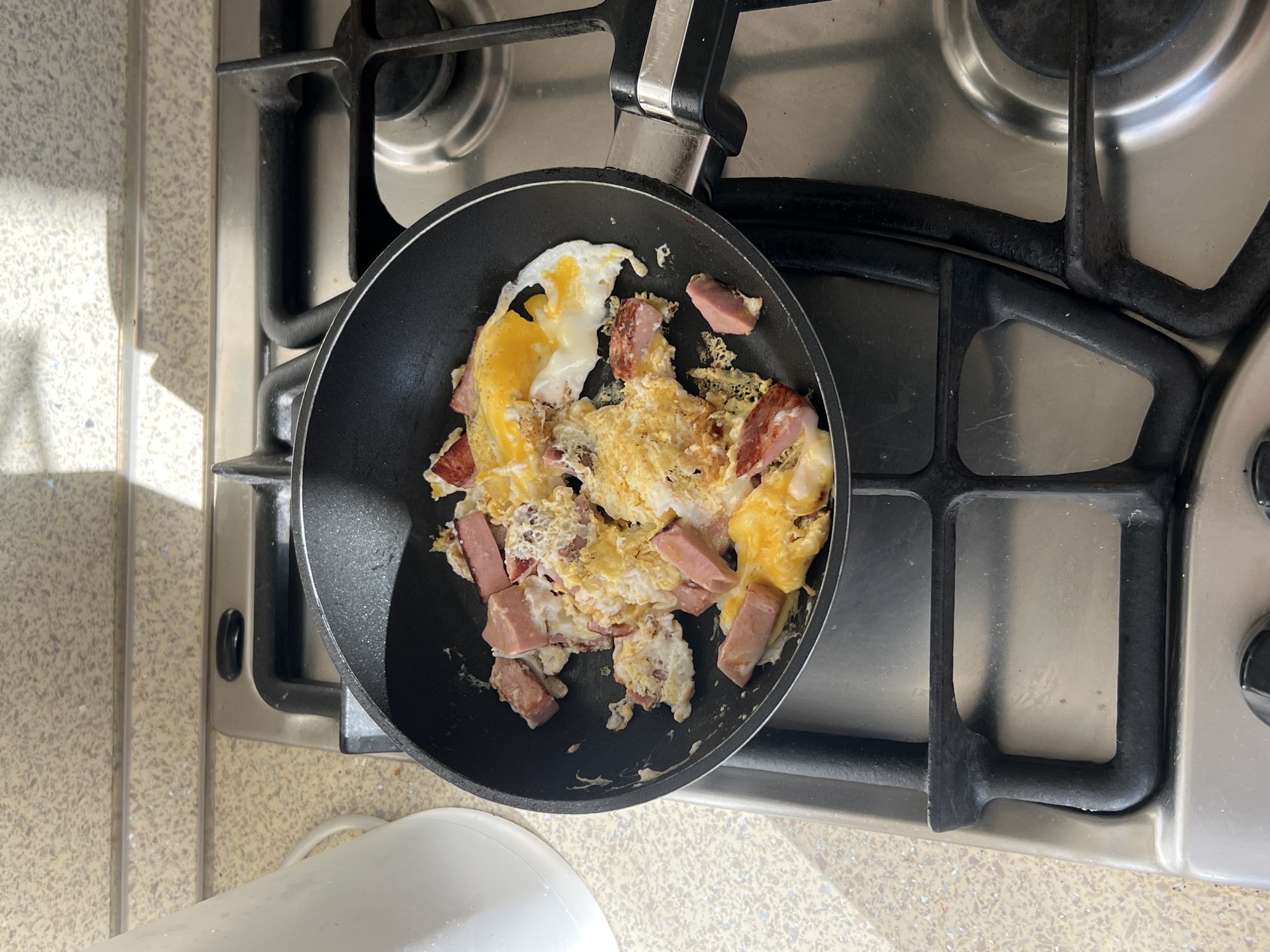 A black frying pan sits on a stainless steel gas stovetop. The pan contains scrambled eggs and pieces of what appears to be sliced sausage or ham. The stovetop has four burners and is speckled with some minor cooking residue. A white, partially visible object is near the bottom left corner of the image, likely part of the kitchen counter. There is no indication of this image being taken in St. Petersburg, Russia.