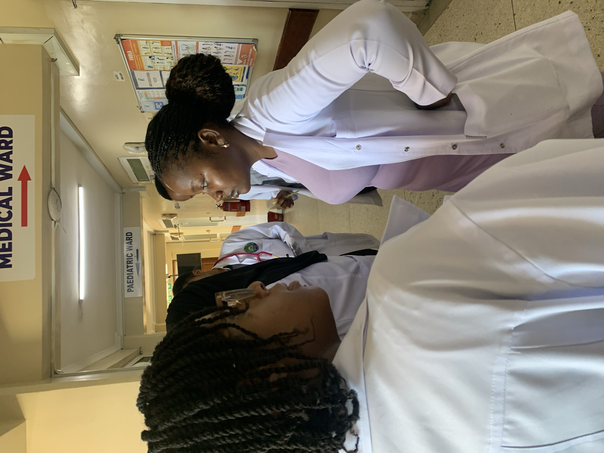 **Location:** Interior of a medical facility, plausibly a hospital, in Dar es Salaam, Tanzania (based on the signs and apparent attire).
**People:** Two African women in medical attire. One is standing, with her hair up in a bun, wearing a white coat and a light purple top. She is looking down and appears to be examining or talking to the second woman, who is lying down wearing glasses and a white coat.
**Objects:** Medical ward sign, Pediatric ward sign, hospital hallway, informational poster regarding Ebola, fire extinguisher, stethoscope.
**Actions:** One woman is examining another.
**Interactions:** A medical professional examining a patient.
**Environment:** Hospital hallway, fluorescent lighting, signs indicating medical wards.