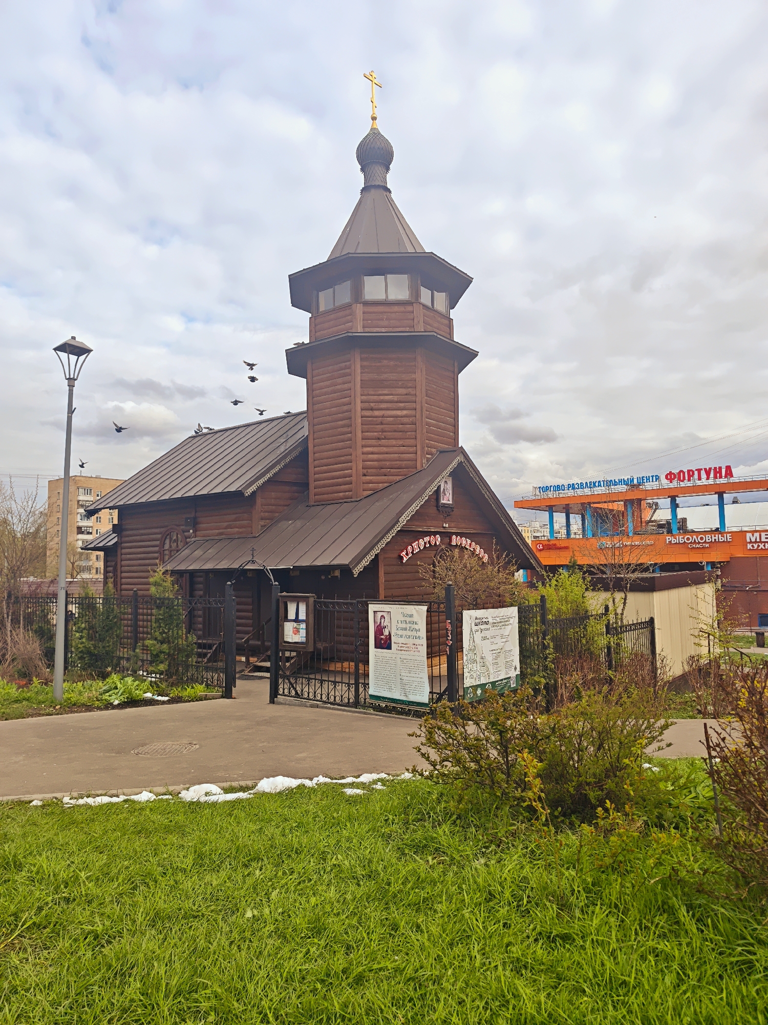 An outdoor, eye-level shot captures a traditional wooden Orthodox church under an overcast sky in Moscow, Russia. The church, constructed from dark brown logs, features a distinctive octagonal tower topped with a dark dome and a golden Orthodox cross. A wooden entrance porch is visible on the left side of the main structure.
In the foreground, a vibrant green grassy area shows patches of melting snow, suggesting spring. A paved path leads towards the church's metal fence and open gate. A tall grey lamppost stands to the left of the path. Several birds are seen flying above the church roof and perched on it.
Behind the church, a large orange building with red lettering dominates the background. This building is identified by the text "ТОРГОВО-РАЗВЛЕКАТЕЛЬНЫЙ ЦЕНТР ФОРТУНА" (Shopping and Entertainment Center FORTUNA). Below this, smaller signs read "РЫБОЛОВНЫЕ СНАСТИ" (Fishing Tackle) and "МЕБЕЛЬ КУХНИ" (Furniture Kitchens). Further in the background, a multi-story residential building can be seen.
On the church's facade above an entrance, the phrase "ХРИСТОС ВОСКРЕС!" (Christ is Risen!) is visible. Two information boards with Russian text and images are placed near the fence entrance, providing details about the church or religious events. The scene depicts a blend of traditional religious architecture with modern urban infrastructure. The weather is cloudy and bright, indicative of daytime.