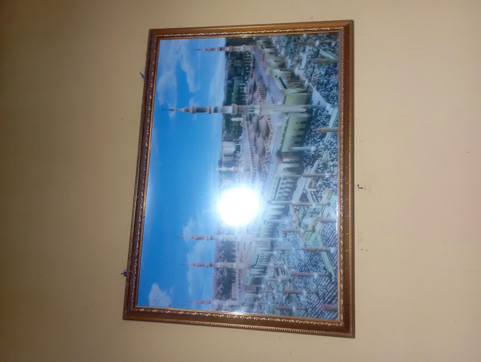 This is a framed photograph of the Prophet's Mosque in Medina, Saudi Arabia, hanging on a plain, light-colored wall. The photograph depicts an aerial view of the mosque complex, showcasing its numerous minarets, domes, and courtyards filled with a large congregation of people. The sunlight in the photograph creates a bright glare in the center, obscuring some details. The frame is ornate and gold-colored. There are small imperfections on the wall around the frame, including what appear to be scuff marks or dirt.