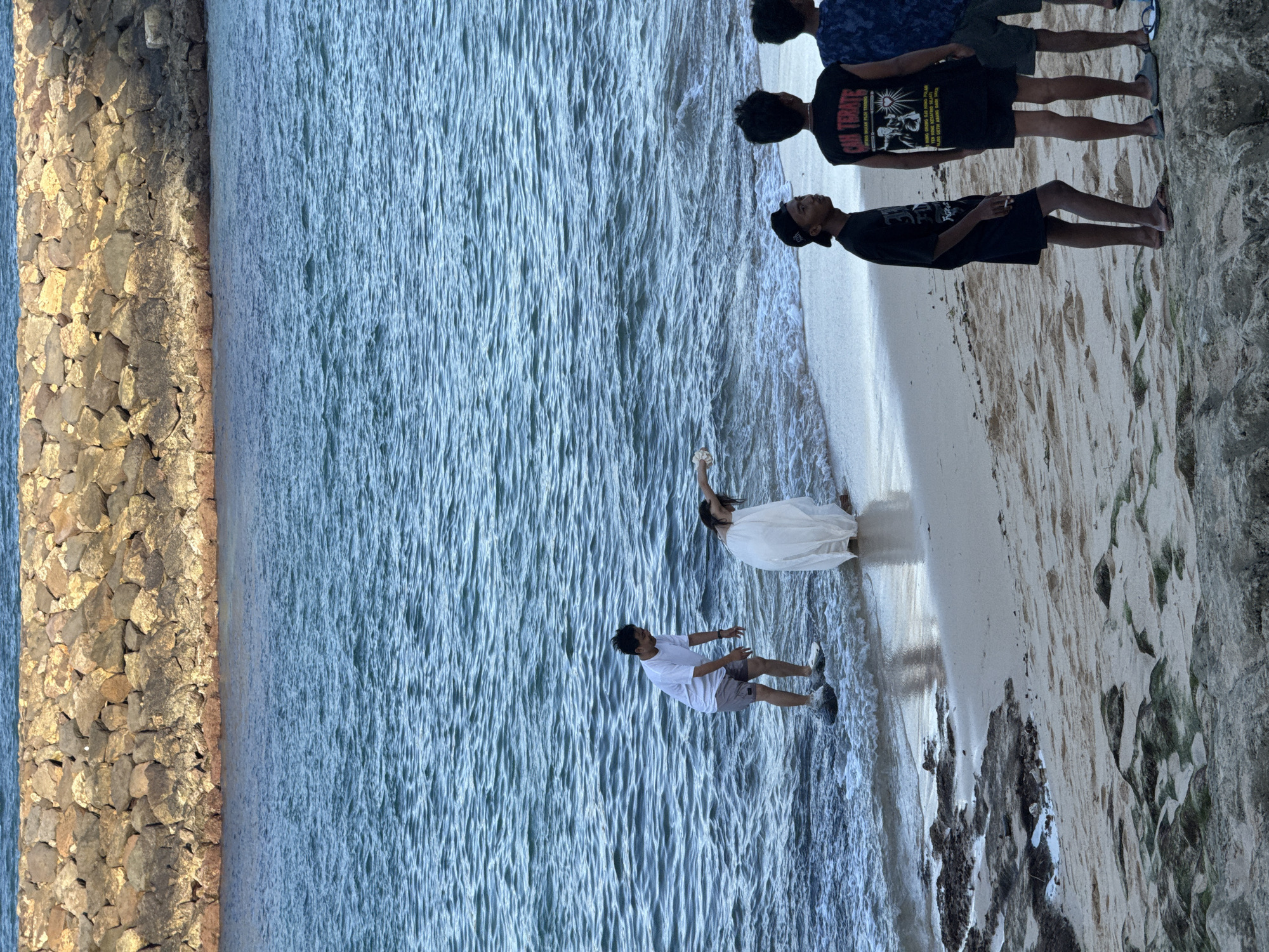 The image depicts a group of individuals on a beach in Kuta Selatan, Indonesia, near a stone breakwater. In the shallow water, a person in a white dress appears to be interacting with another individual in a white t-shirt and shorts, who is standing on a dark object in the water, possibly a surfboard. The person in the dress is bending over, reaching towards the water. On the sandy shore, several young men are gathered, observing the scene. One man in a black t-shirt with "CAH TERATE" printed on it, and another in a blue patterned shirt are visible. The environment includes the ocean with moderate waves, the sandy beach, and a rocky shoreline. The lighting suggests it is either early morning or late afternoon.